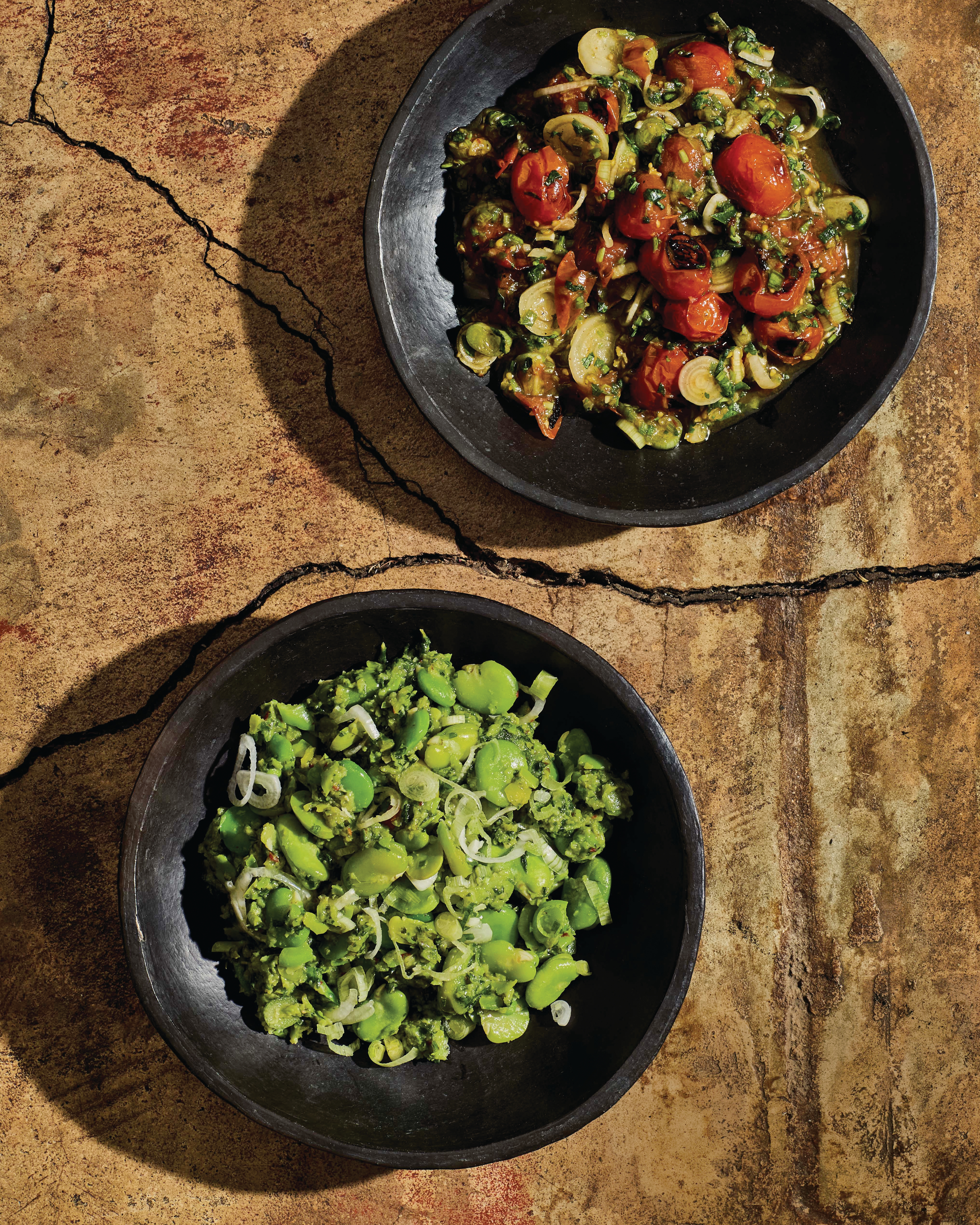 Two black bowls on stone surface - top with roasted tomato vegetable mix, bottom with bright green bean salad.