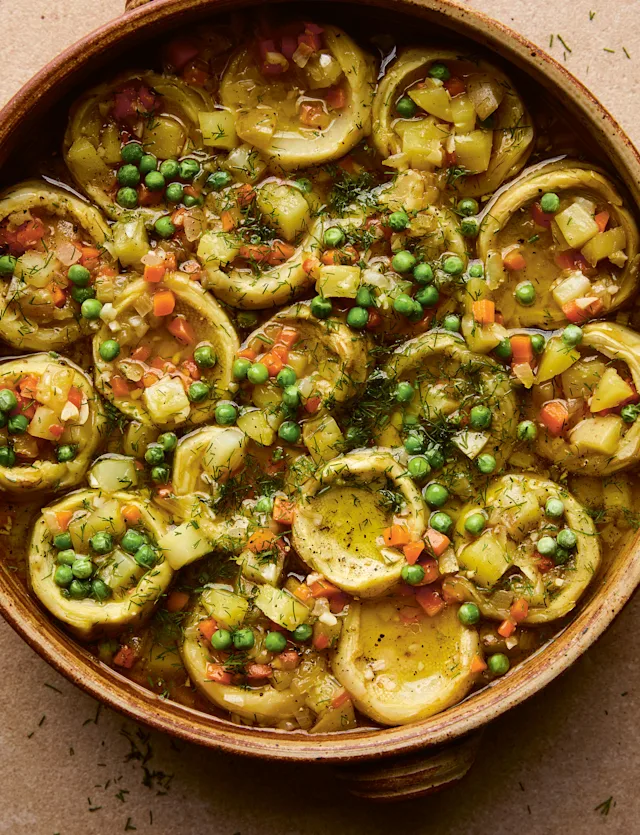 A rustic ceramic bowl filled with braised artichoke hearts, green peas, diced carrots, and fresh dill in a savory broth.