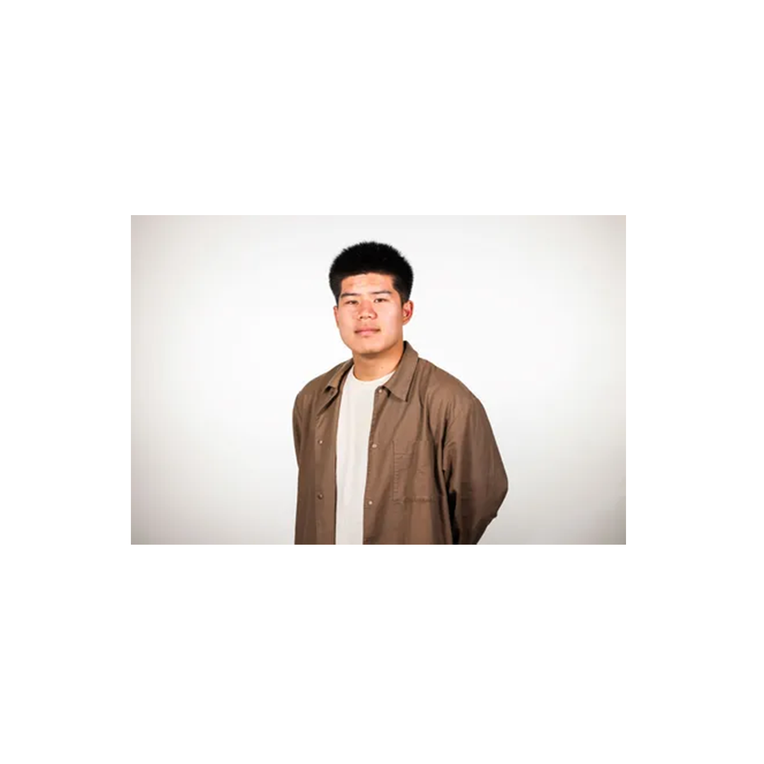 Young person with short dark hair wearing a brown button-up shirt over a white t-shirt against a light background.