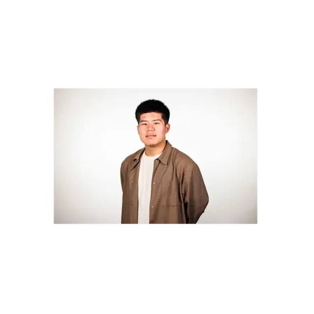 Young person with short dark hair wearing a brown button-up shirt over a white t-shirt against a light background.