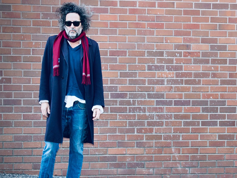 Person with wild hair wearing sunglasses, navy coat, red scarf and jeans standing against a brick wall.