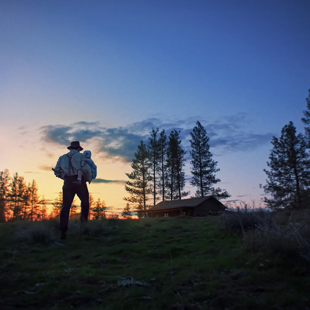 Hiker with backpack walking toward cabin at sunset, silhouetted against blue sky with pine trees surrounding the area.
