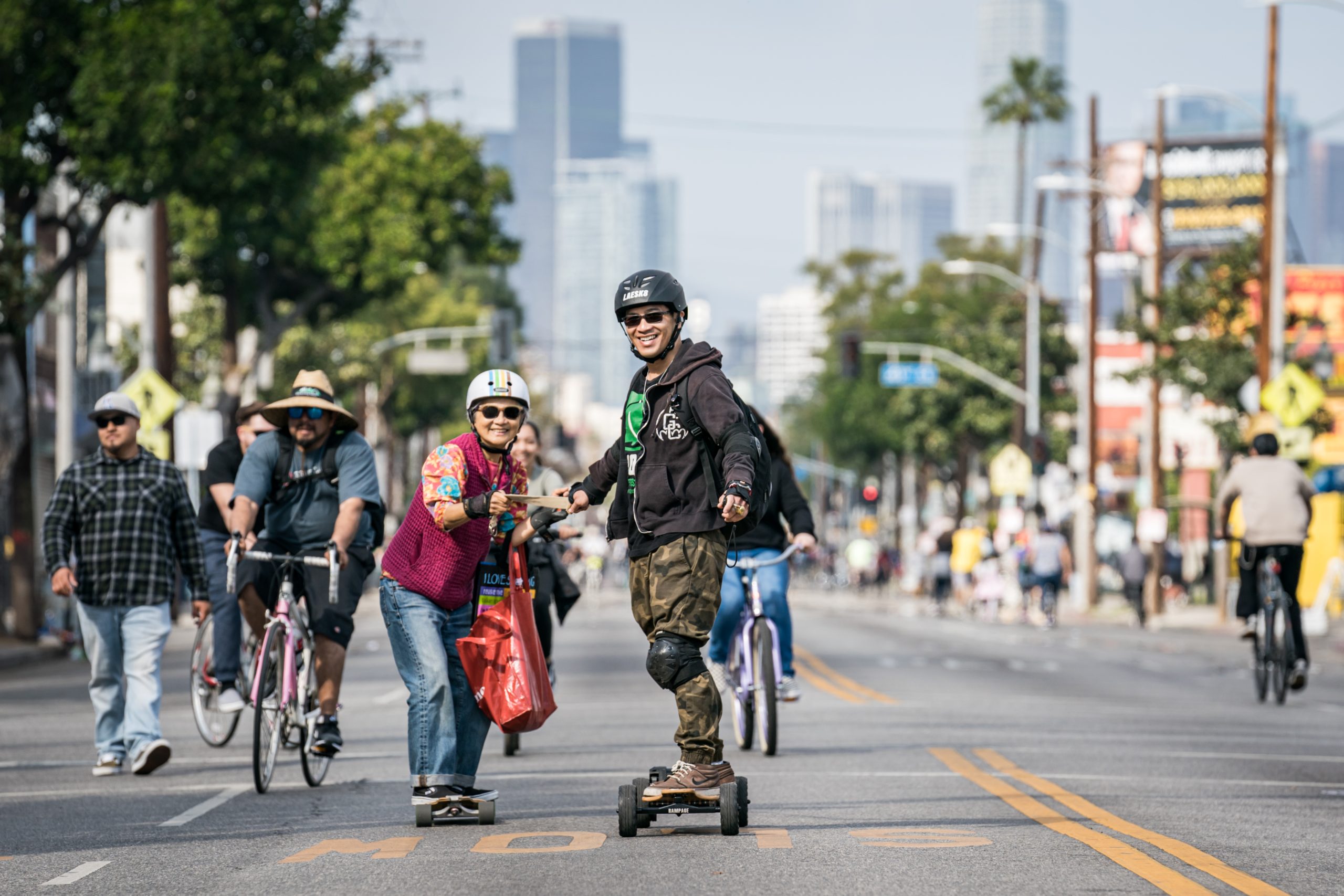 Two people skateboard down a car-free Los Angeles street surrounded by cyclists and pedestrians, with the city skyline in the background.