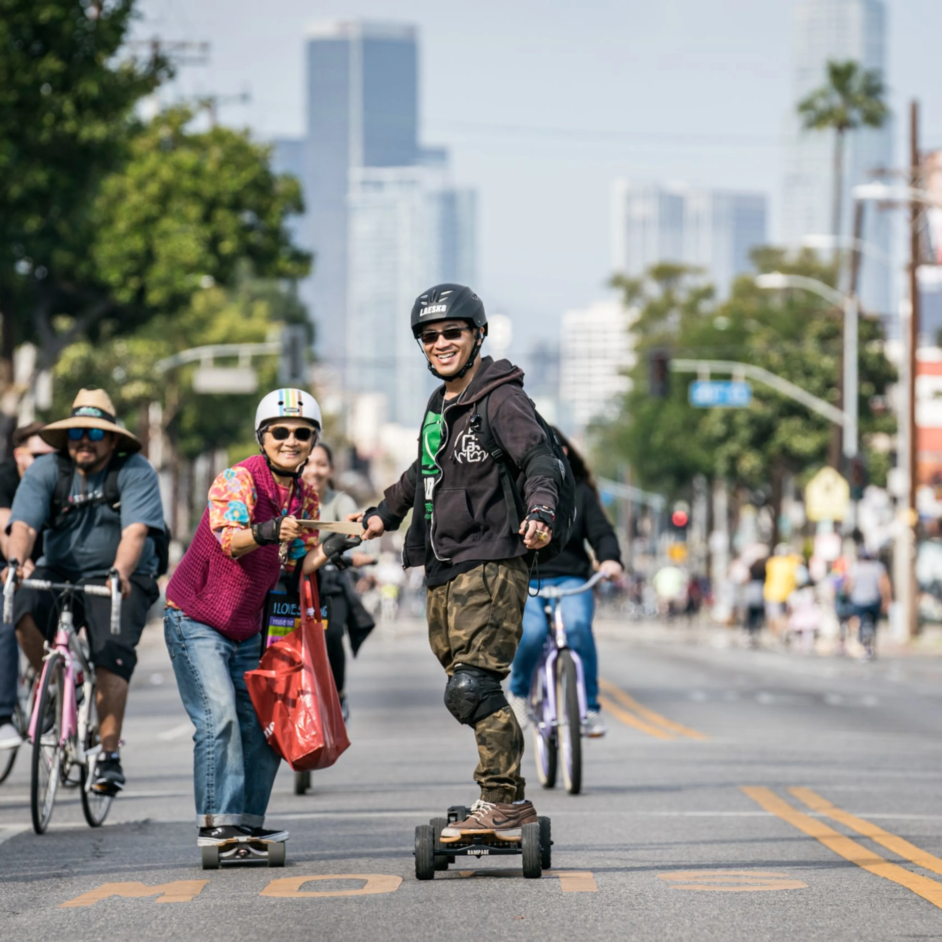 Two people skateboard down a car-free Los Angeles street surrounded by cyclists and pedestrians, with the city skyline in the background.