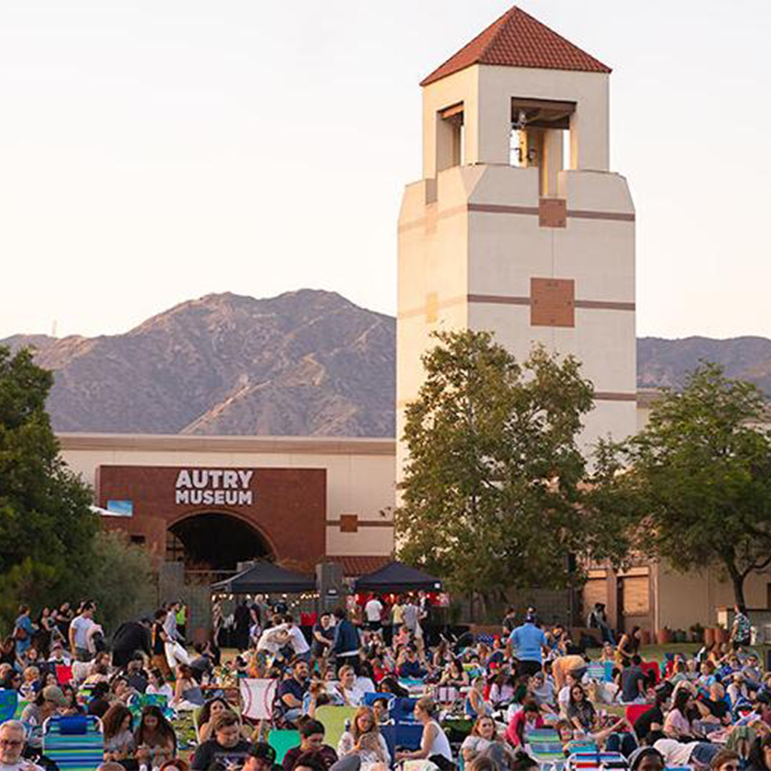 Crowd gathered for outdoor event at Autry Museum with tall bell tower and mountains in background at sunset.