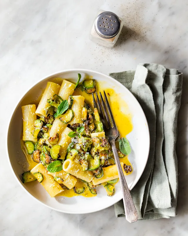 A white bowl of rigatoni pasta with sautéed zucchini, yellow sauce, mint, and Parmesan, served with a fork and green napkin.