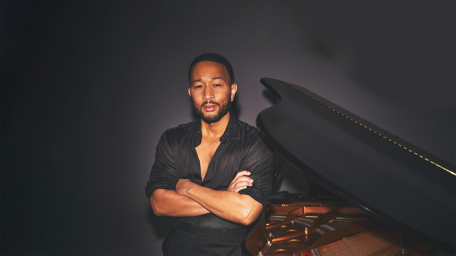 Musician in black shirt standing with arms crossed beside a grand piano against dark background.