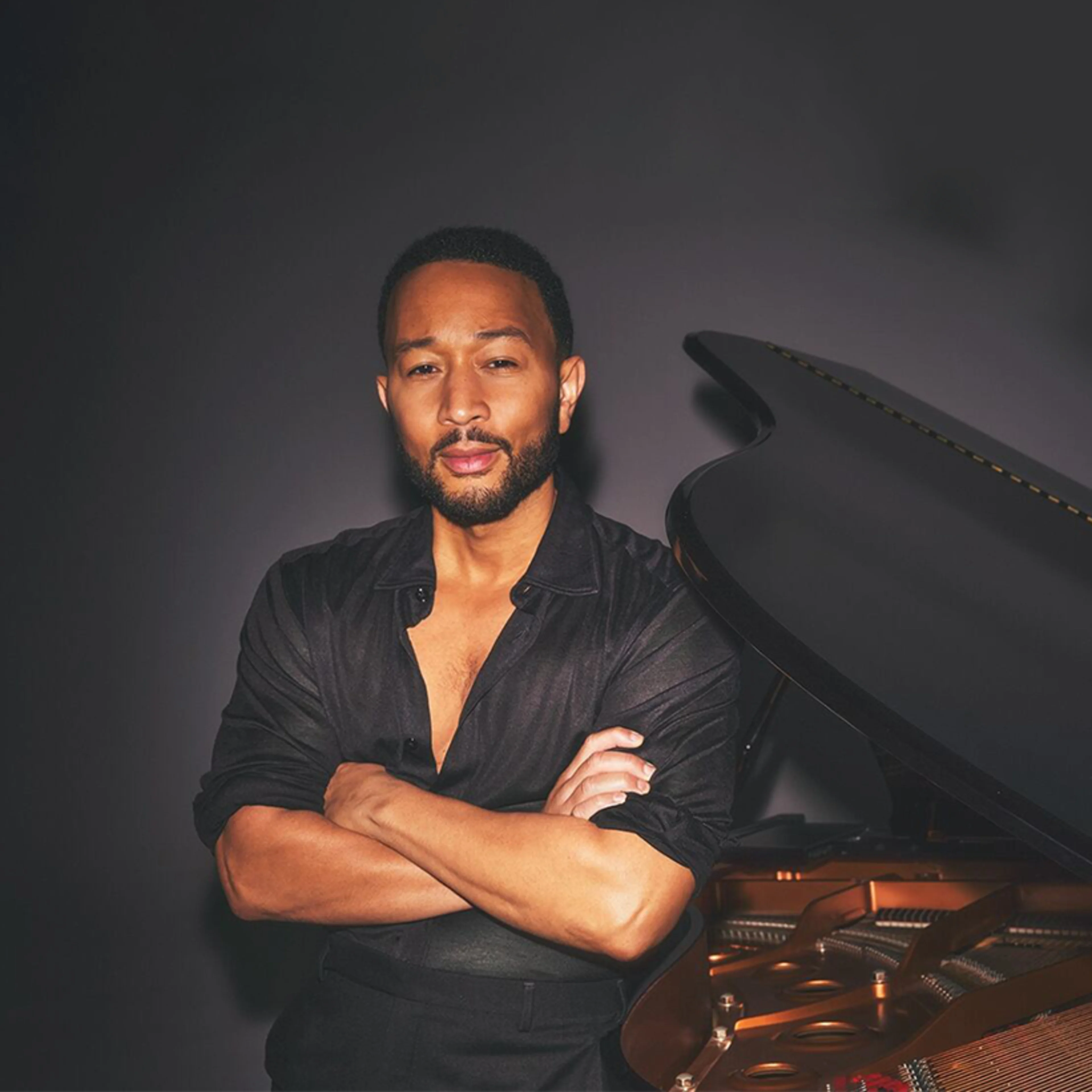 Musician in black shirt standing with arms crossed beside a grand piano against dark background.