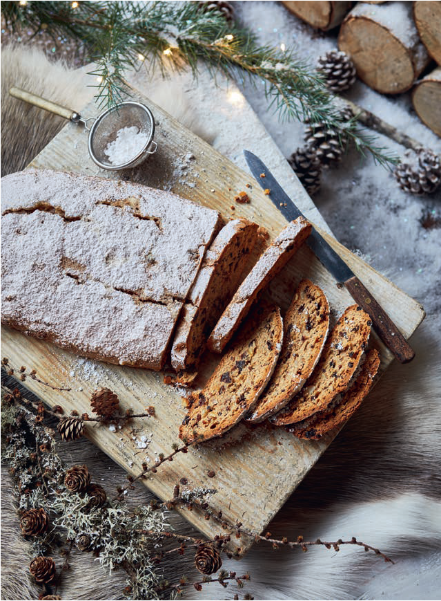 Sliced fruit cake dusted with powdered sugar on wooden board with pine branches and pinecones in winter setting.