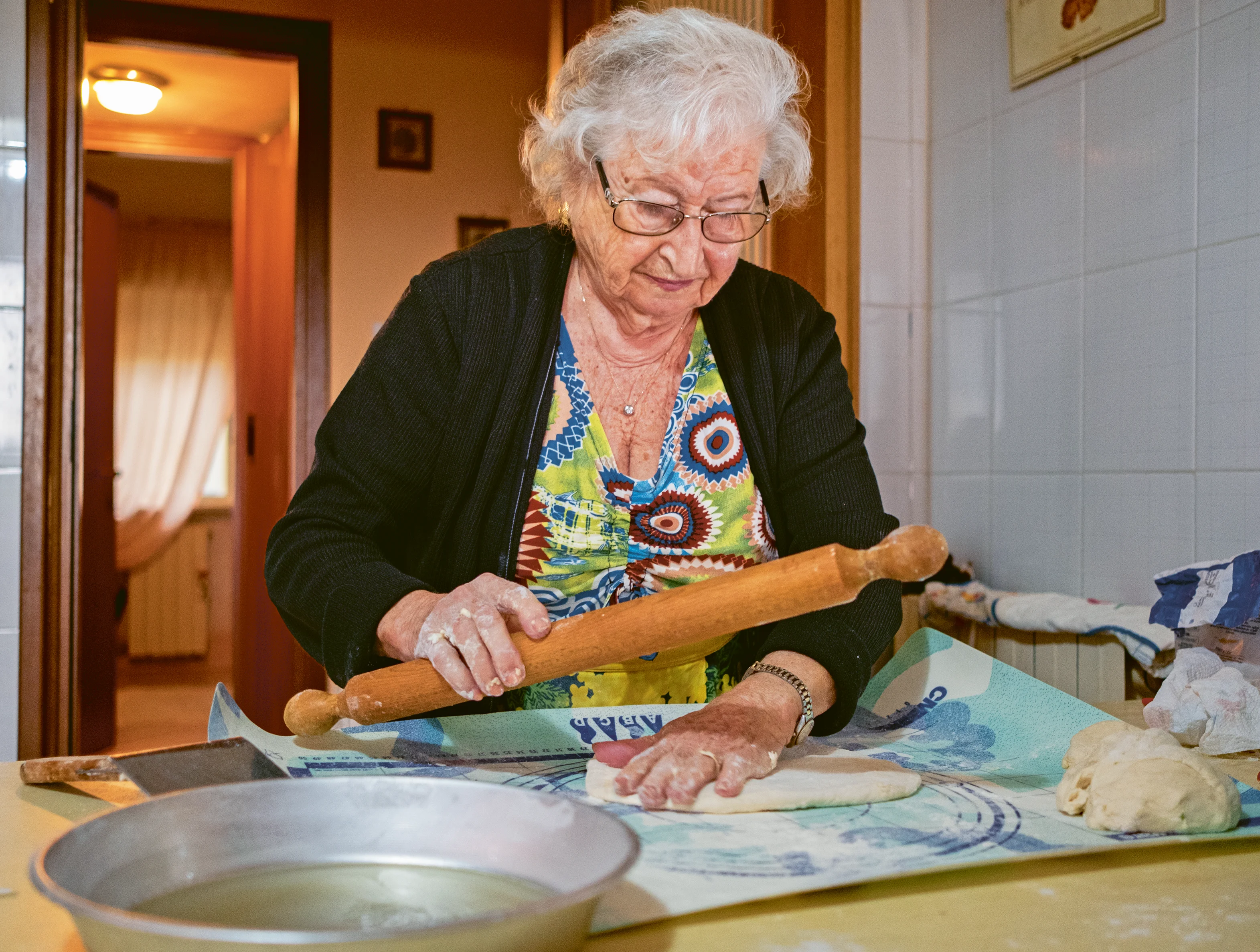 An elderly woman with white hair and glasses rolls dough with a wooden rolling pin on a kitchen table.