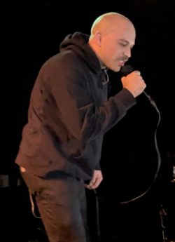 A performer in a dark hoodie singing into a microphone on stage against a black background.