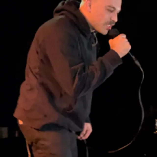 A performer in a dark hoodie singing into a microphone on stage against a black background.