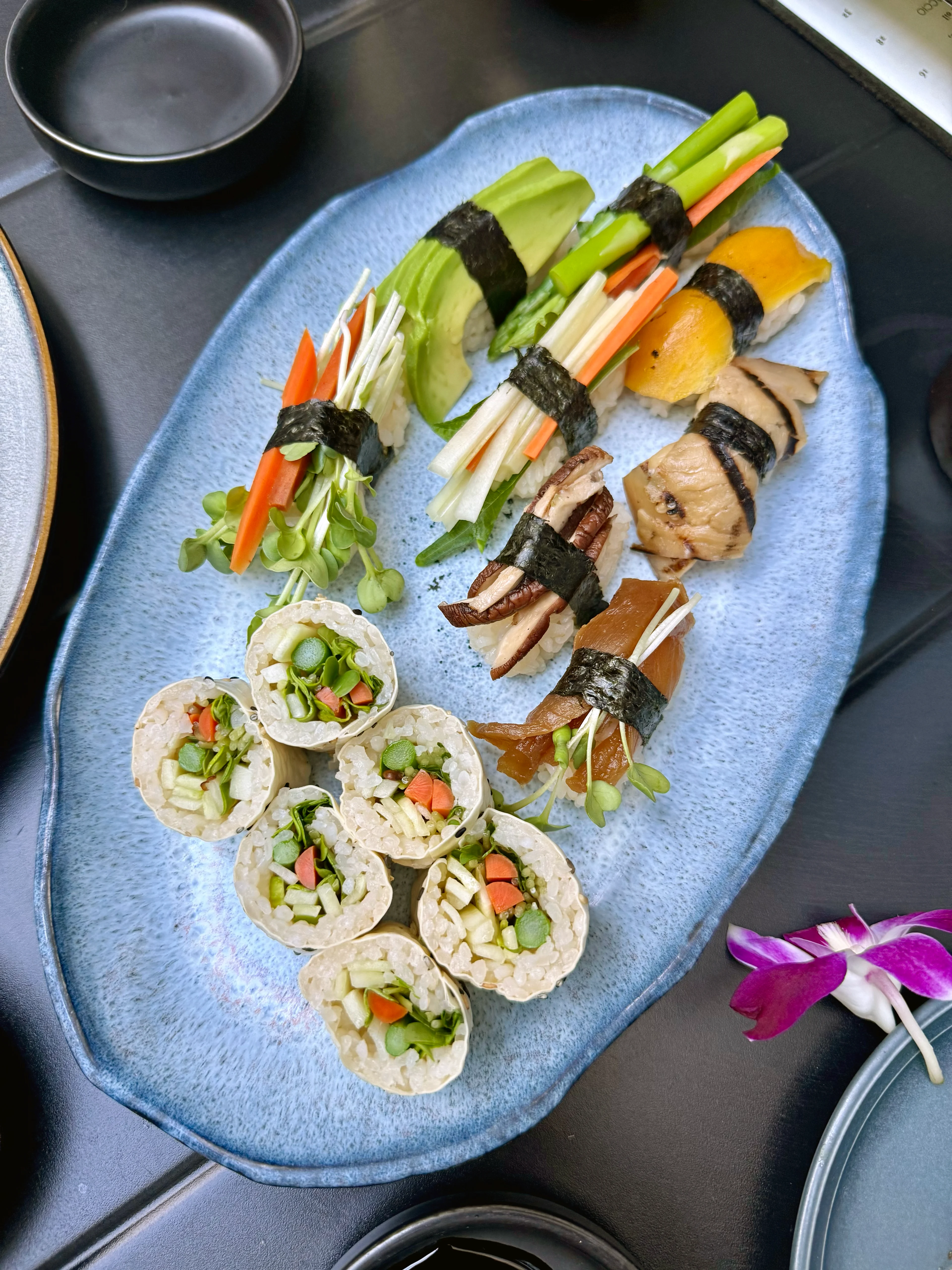 A blue oval plate with assorted vegetable sushi rolls and nigiri topped with colorful veggies, mushrooms, and avocado.