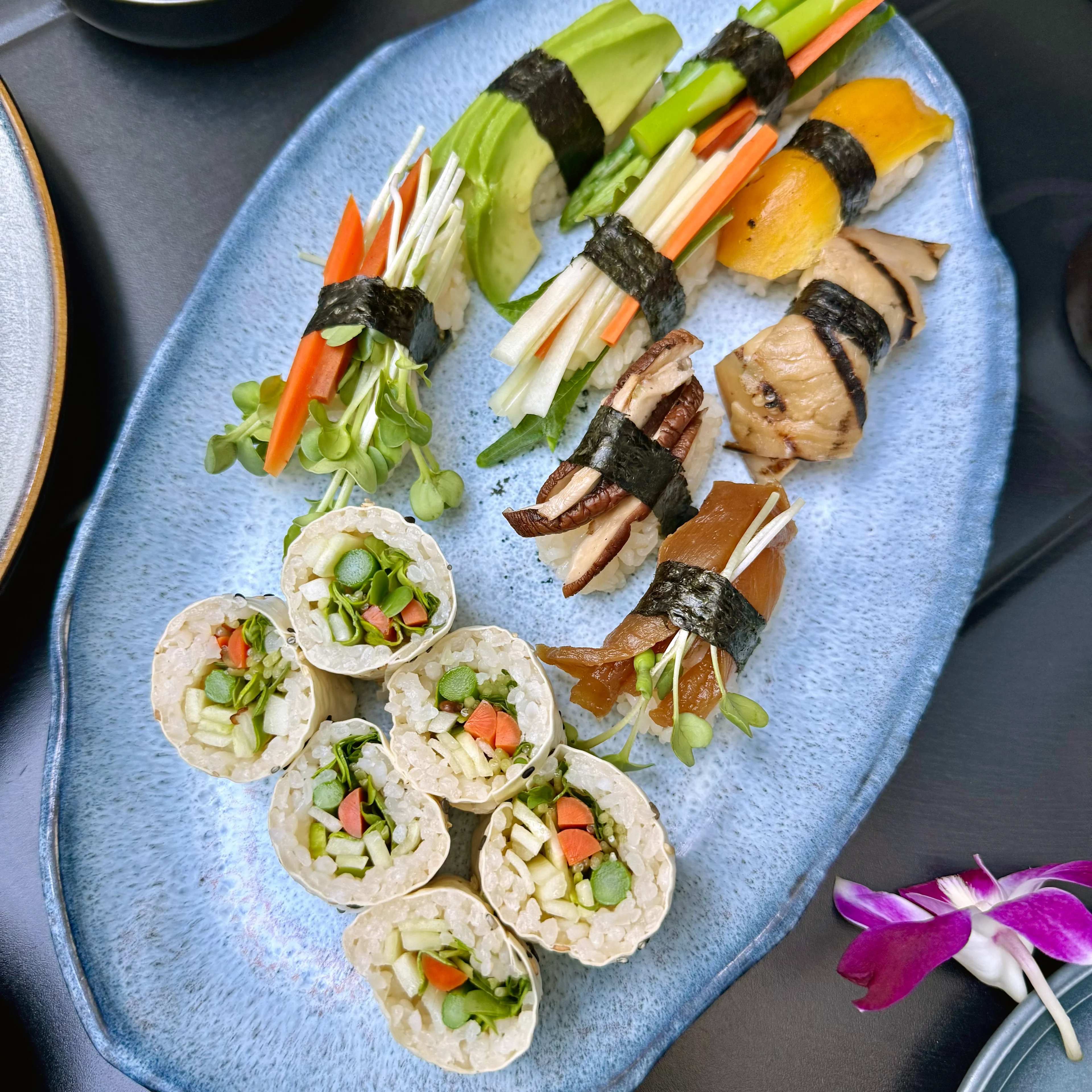 A blue oval plate with assorted vegetable sushi rolls and nigiri topped with colorful veggies, mushrooms, and avocado.