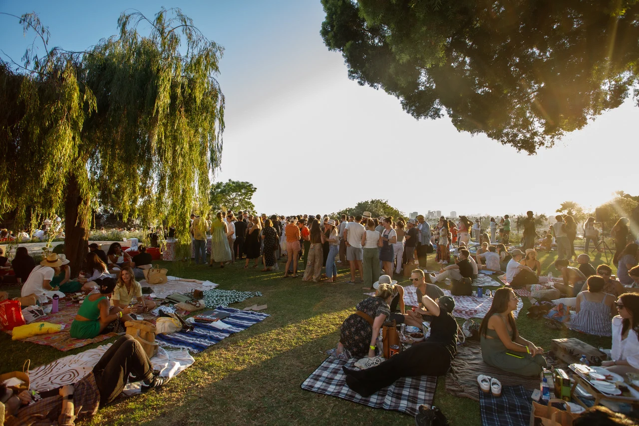 A large crowd gathers in a sunny park, with people sitting on blankets and mingling near willow trees at Barnsdall Fridays Wine Night