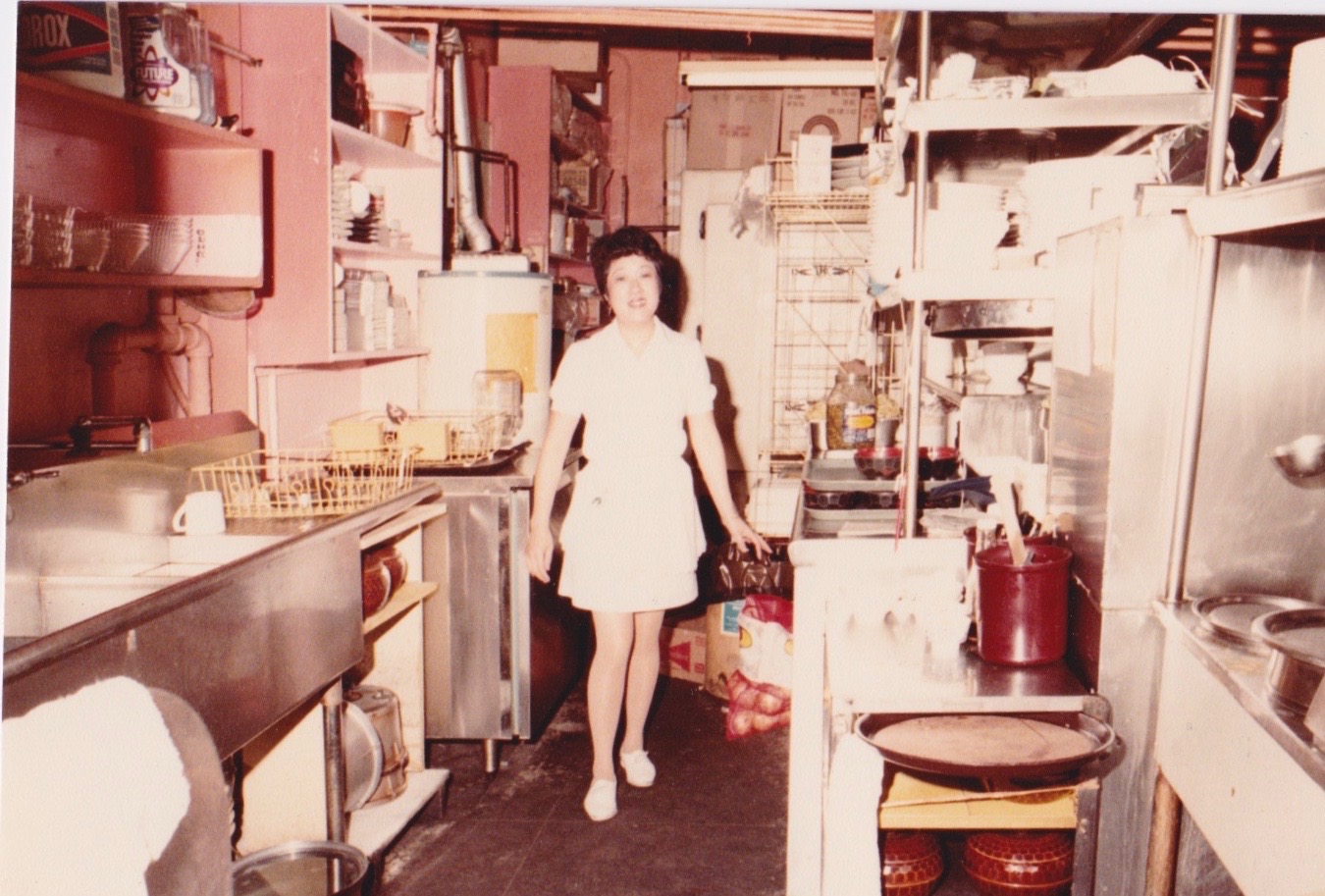 A woman in a white dress stands smiling in a cluttered commercial kitchen with shelves, pots, and equipment surrounding her.