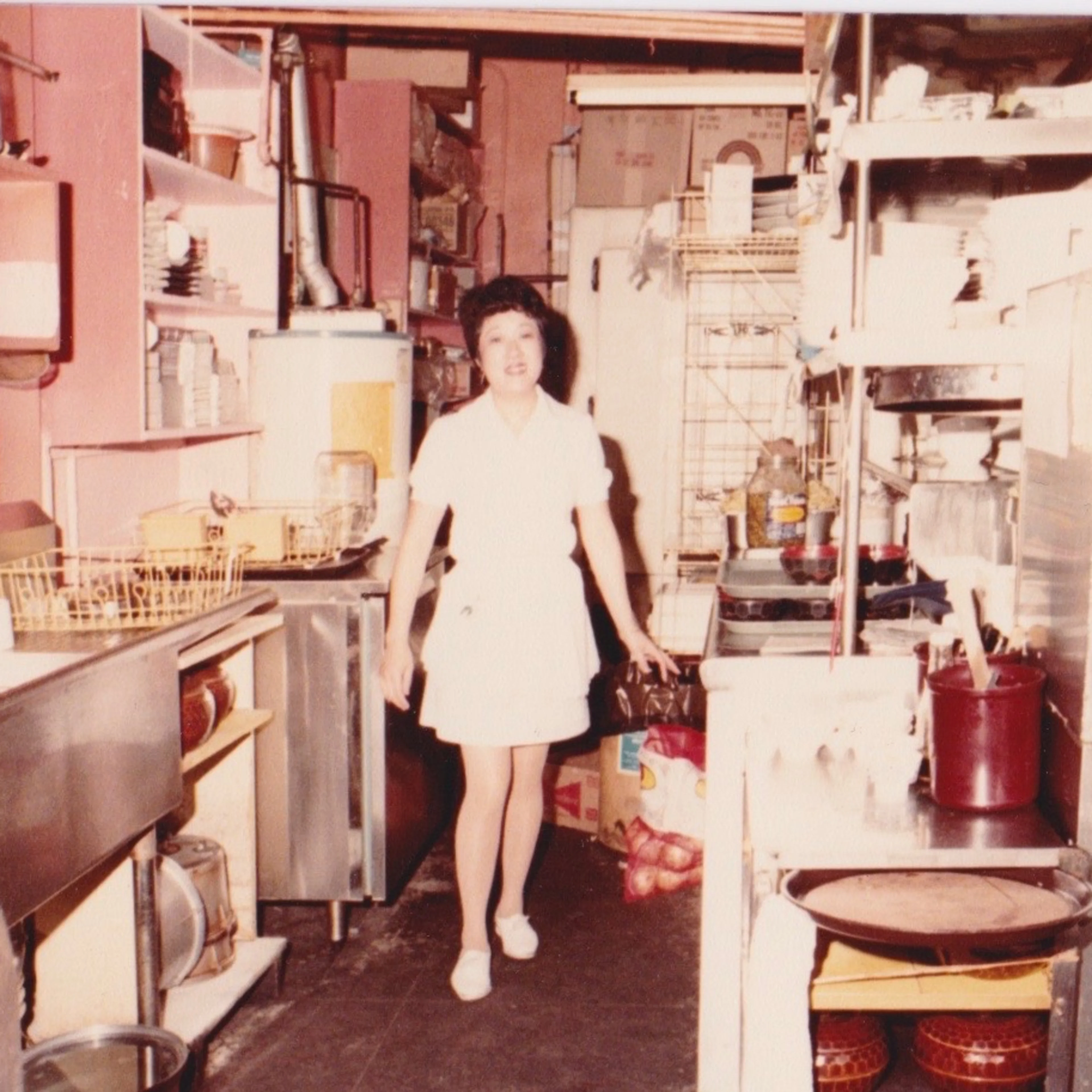A woman in a white dress stands smiling in a cluttered commercial kitchen with shelves, pots, and equipment surrounding her.
