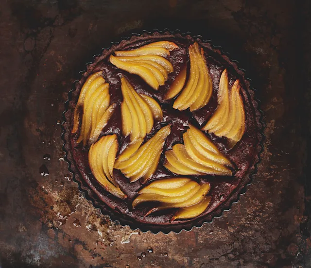 Chocolate tart with fan-arranged poached pear slices on top, served on a rustic dark brown surface.