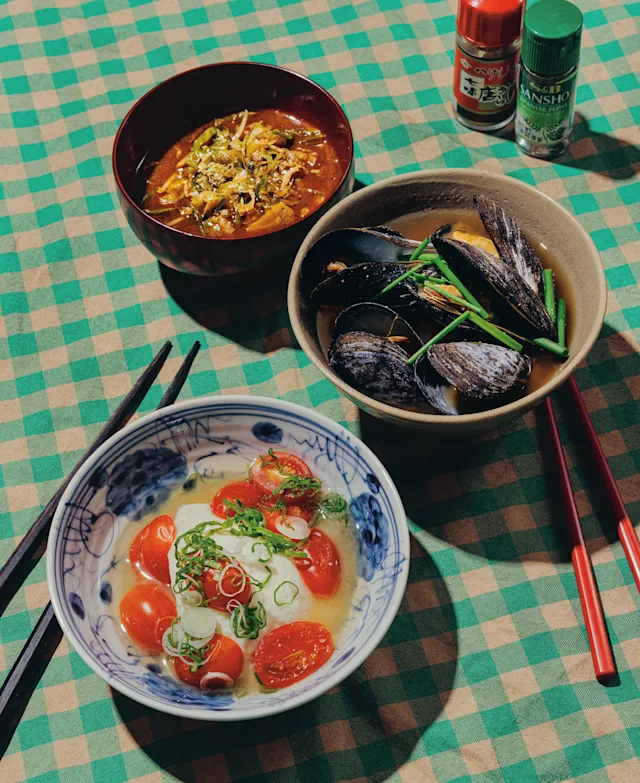Three Asian dishes on a green checkered tablecloth: tomato tofu soup, mussels, and spicy broth, with chopsticks and spices nearby.