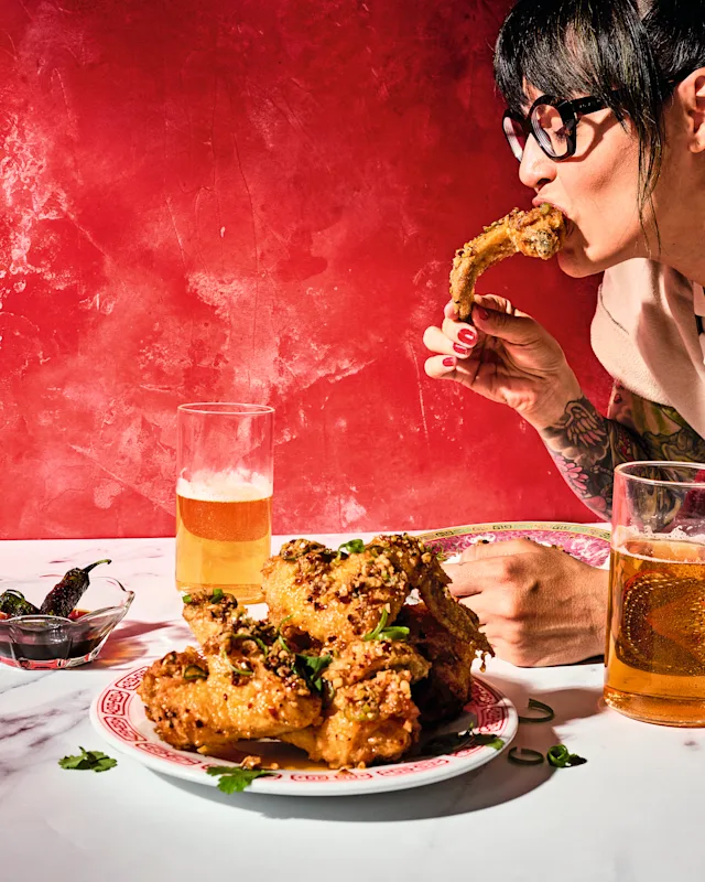 A tattooed woman with glasses eats a chicken wing from a plate of wings, with two glasses of beer and peppers on a marble table.