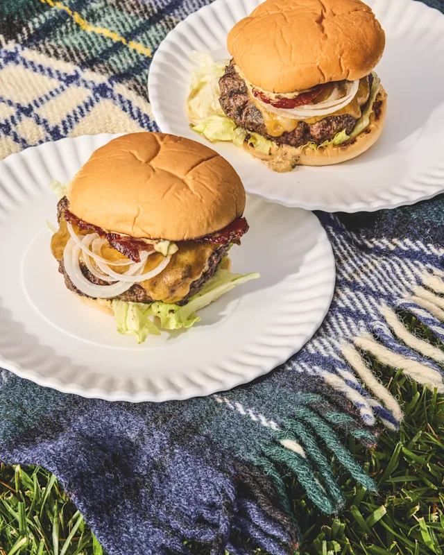 Two loaded cheeseburgers with lettuce, onion, and bacon on white plates atop a plaid blanket on grass.