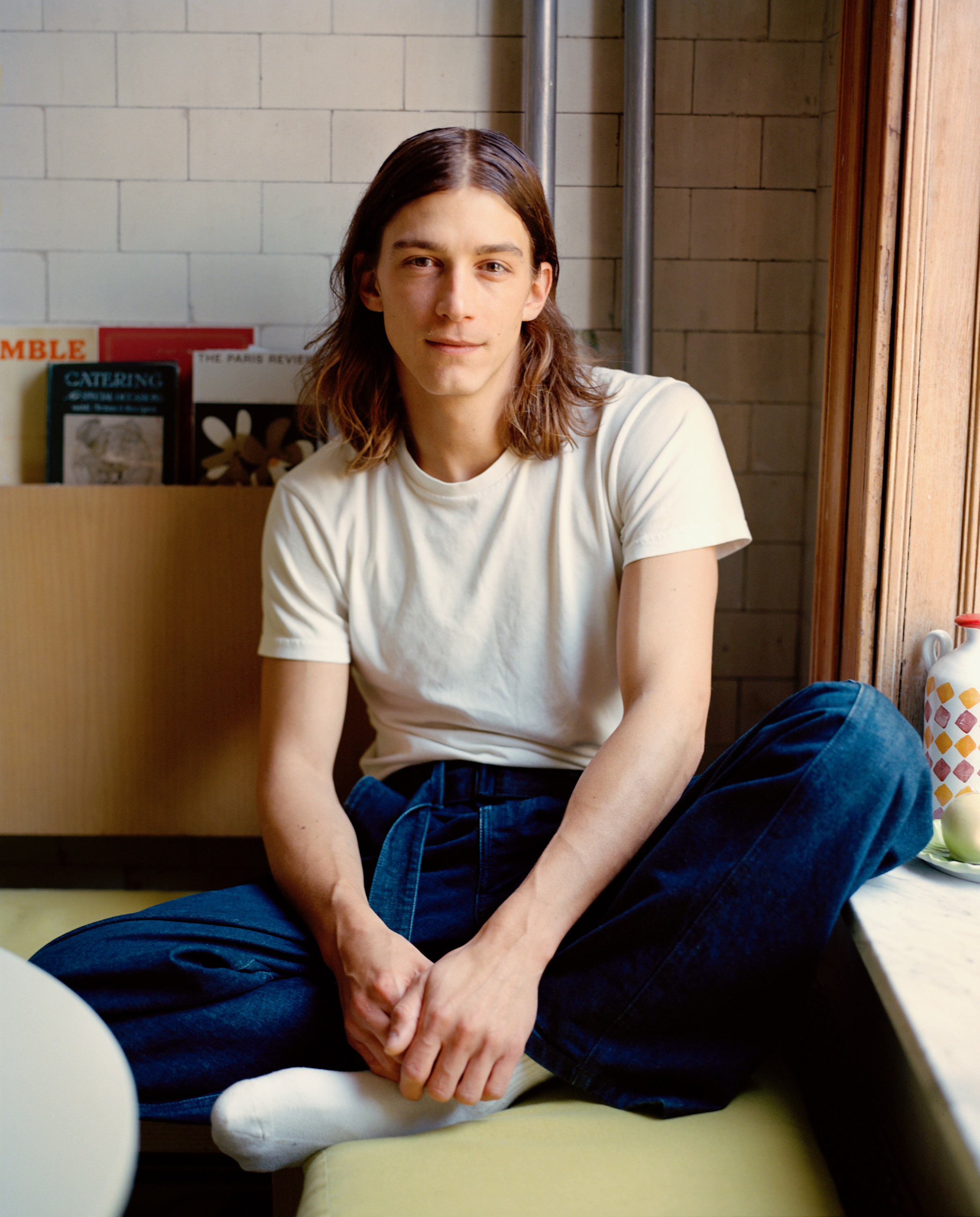 A young person with long brown hair sits cross-legged in a cafe, wearing a white t-shirt and blue jeans.