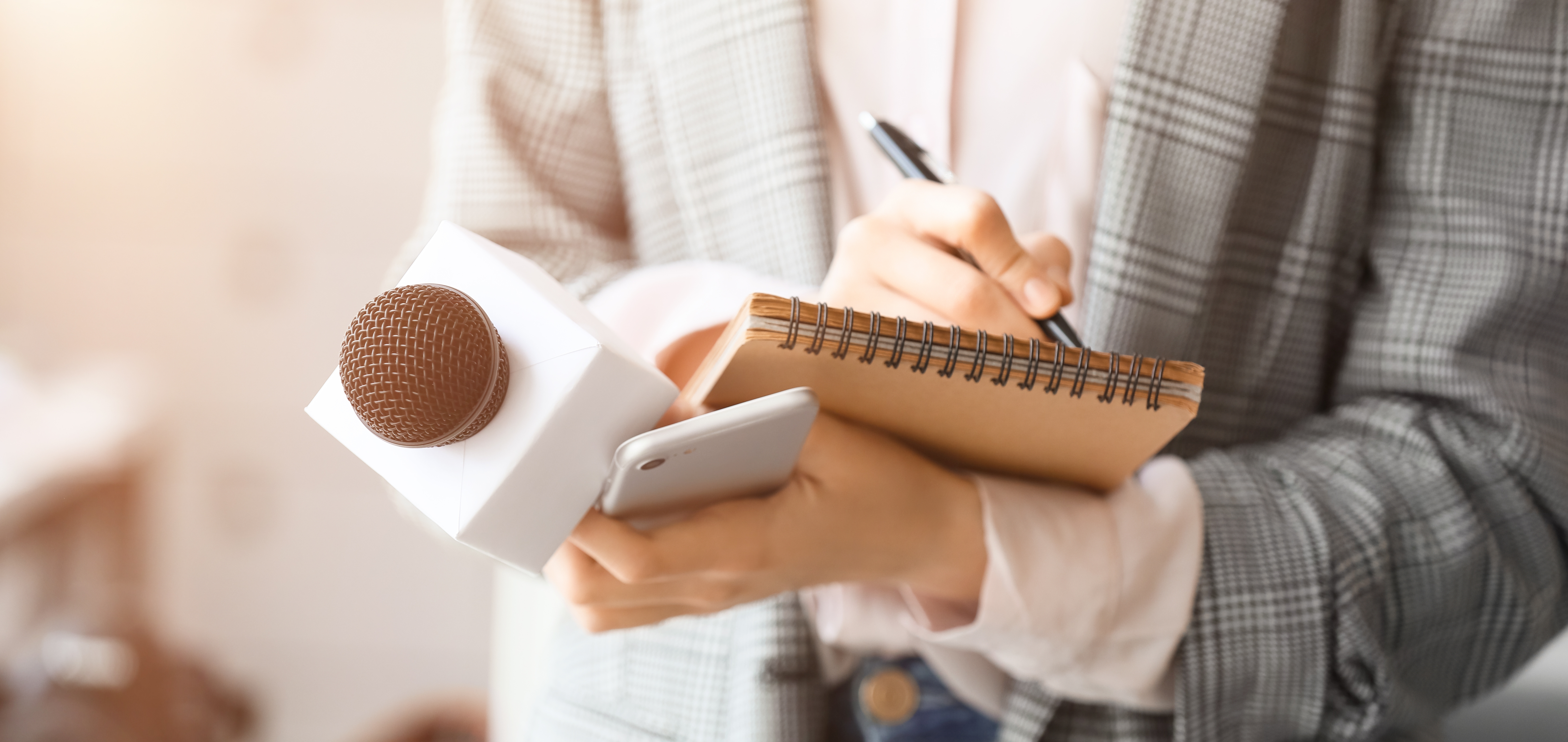 Person in plaid outfit taking notes in spiral notebook while holding smartphone and microphone for interview.