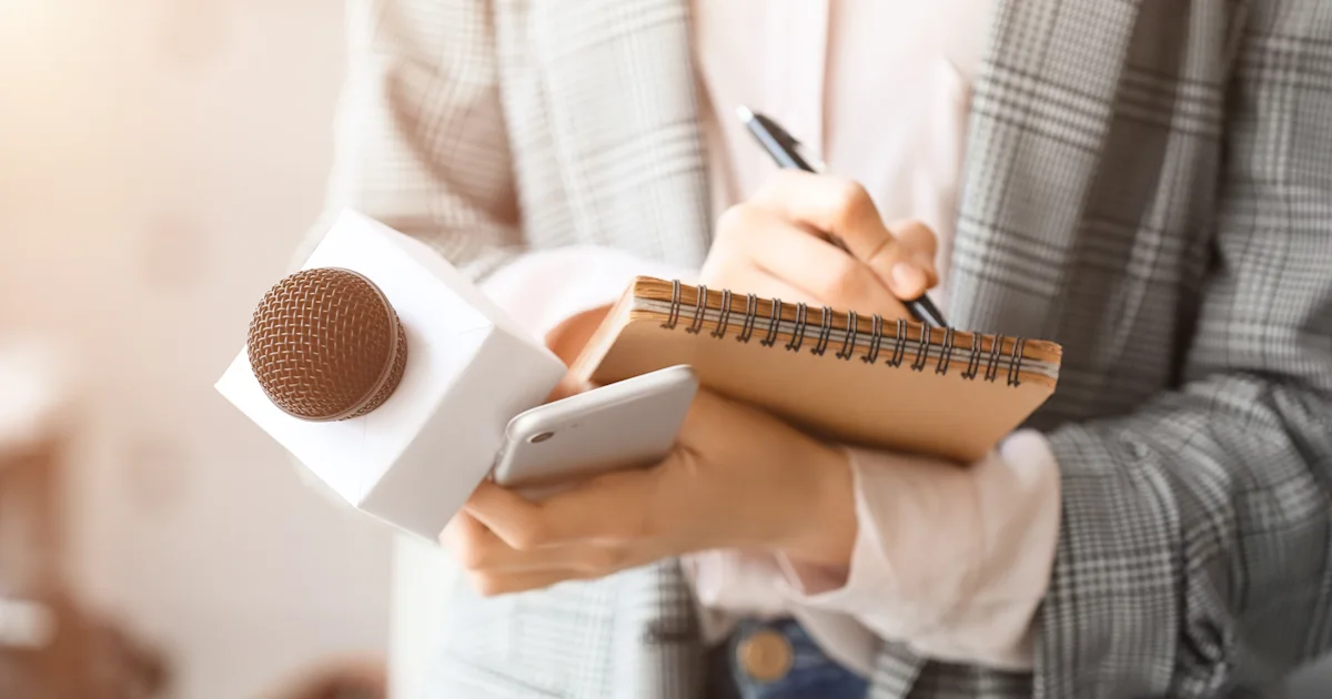 Person in plaid outfit taking notes in spiral notebook while holding smartphone and microphone for interview.