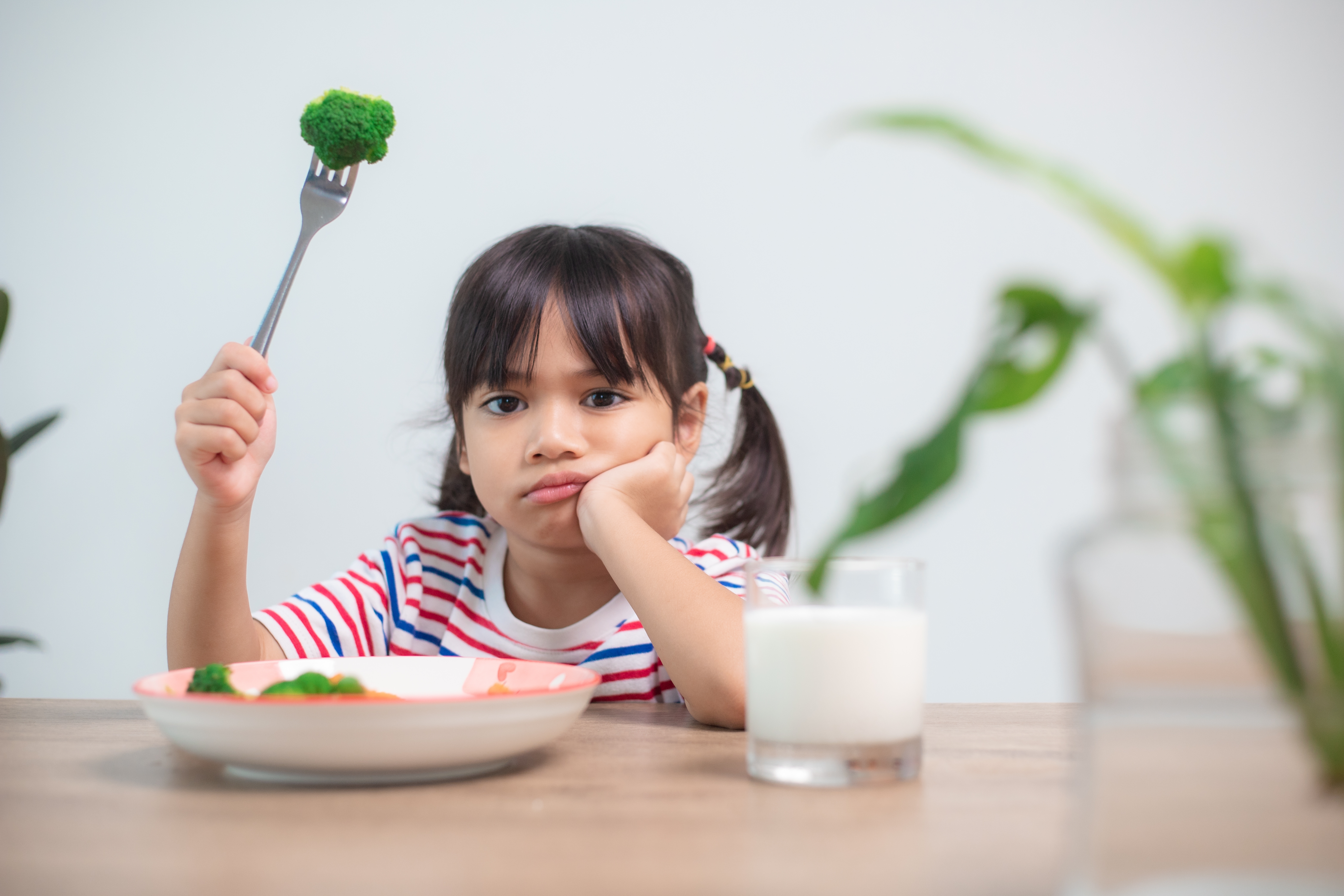 Child in striped shirt reluctantly holding broccoli on fork, looking unenthusiastic about eating vegetables.