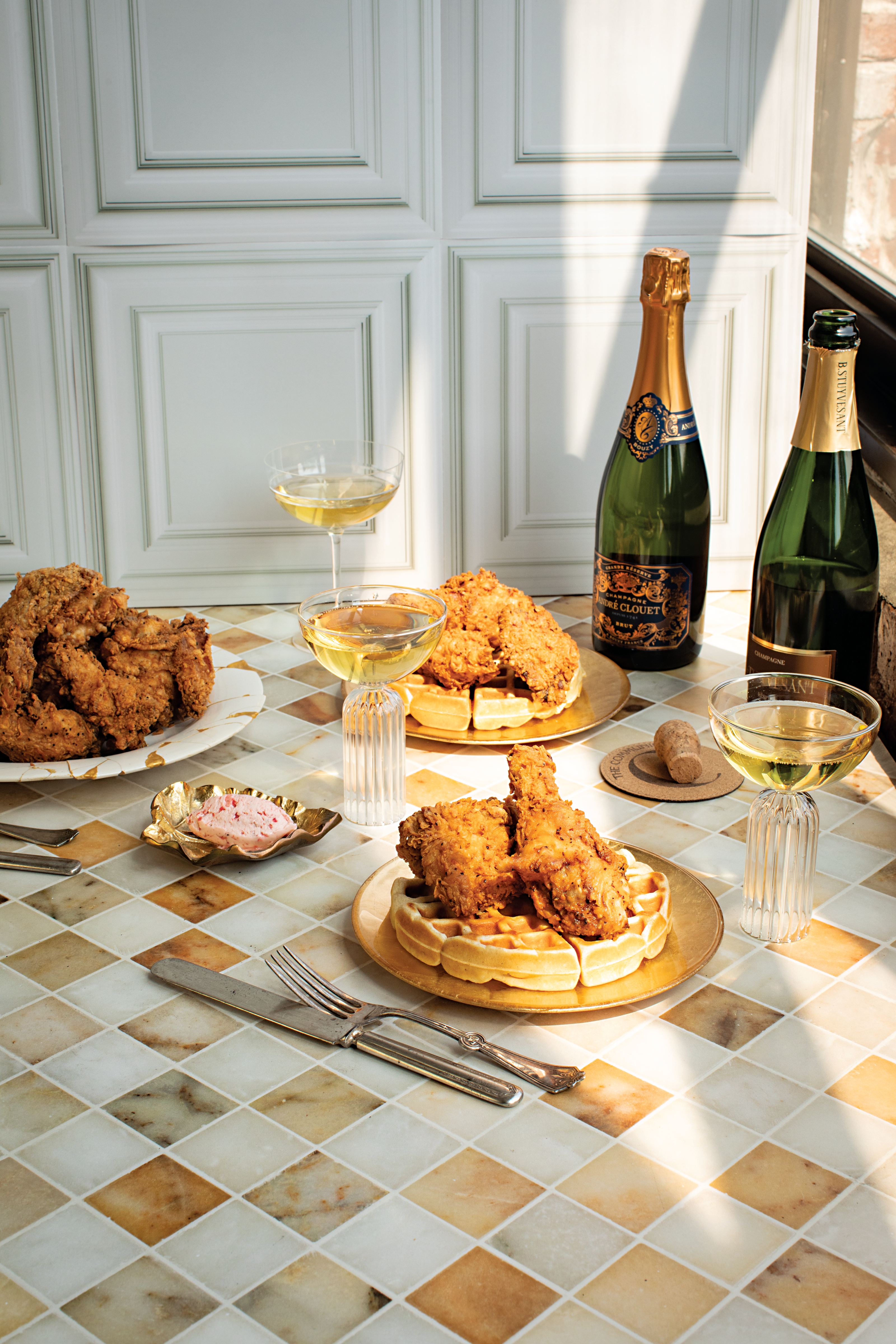 Elegant table setting with fried chicken and waffles, champagne glasses, and bottles on a tiled surface in sunlight.