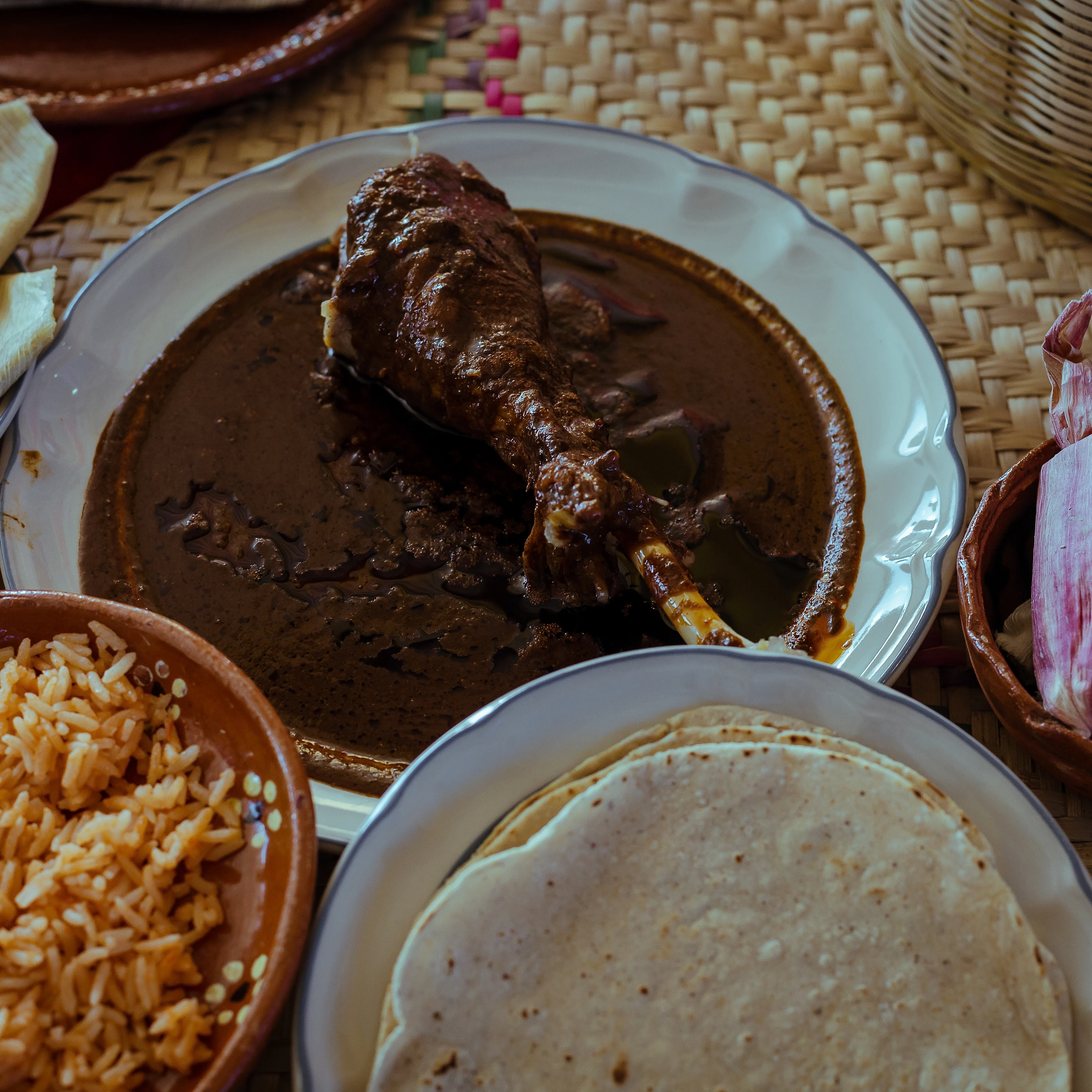 A turkey leg slathered in mole poblano sits on a table along with homemade corn tortillas and red rice.