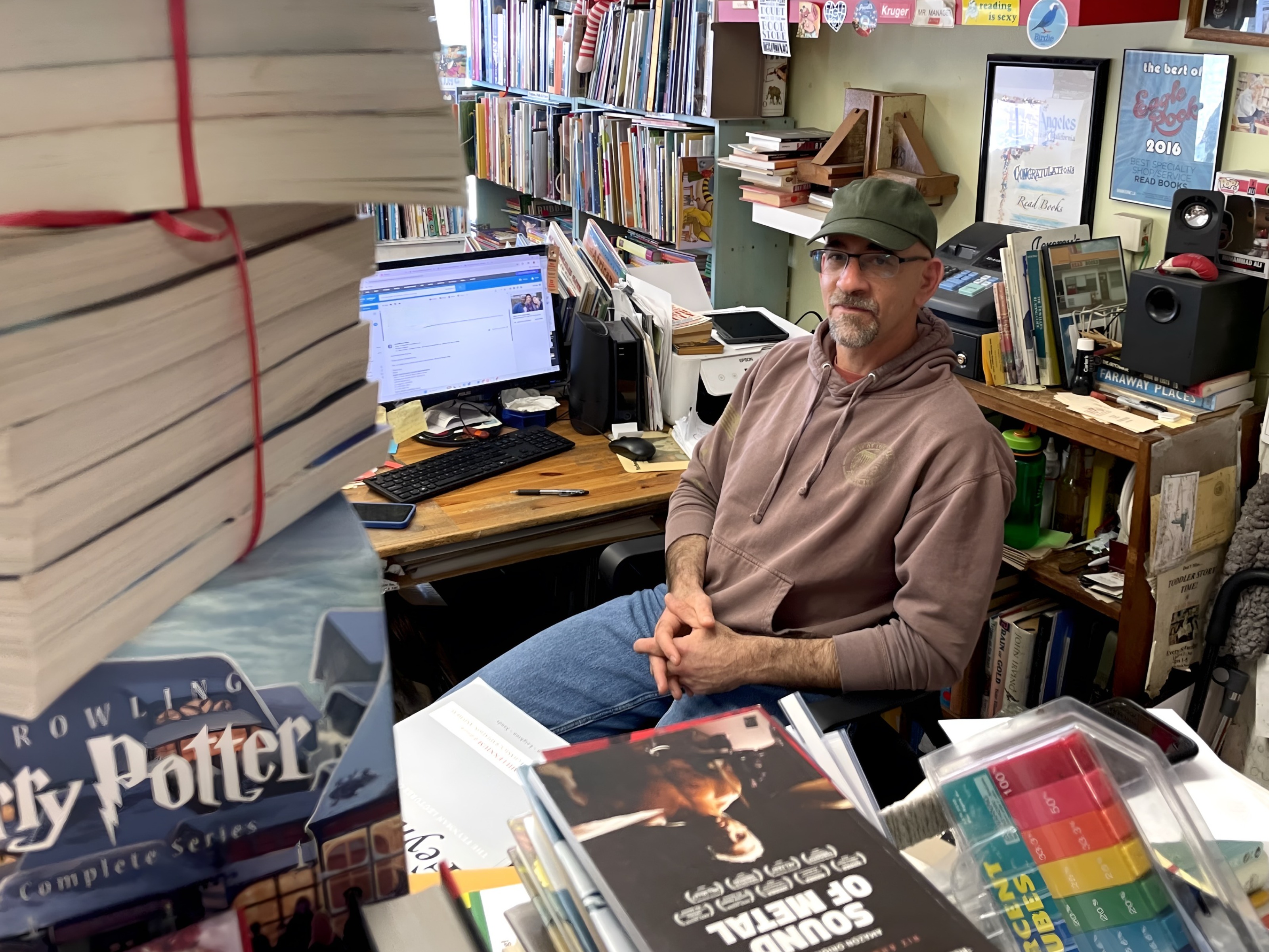 A man sits at a bookstore register surrounded by stacks of books.