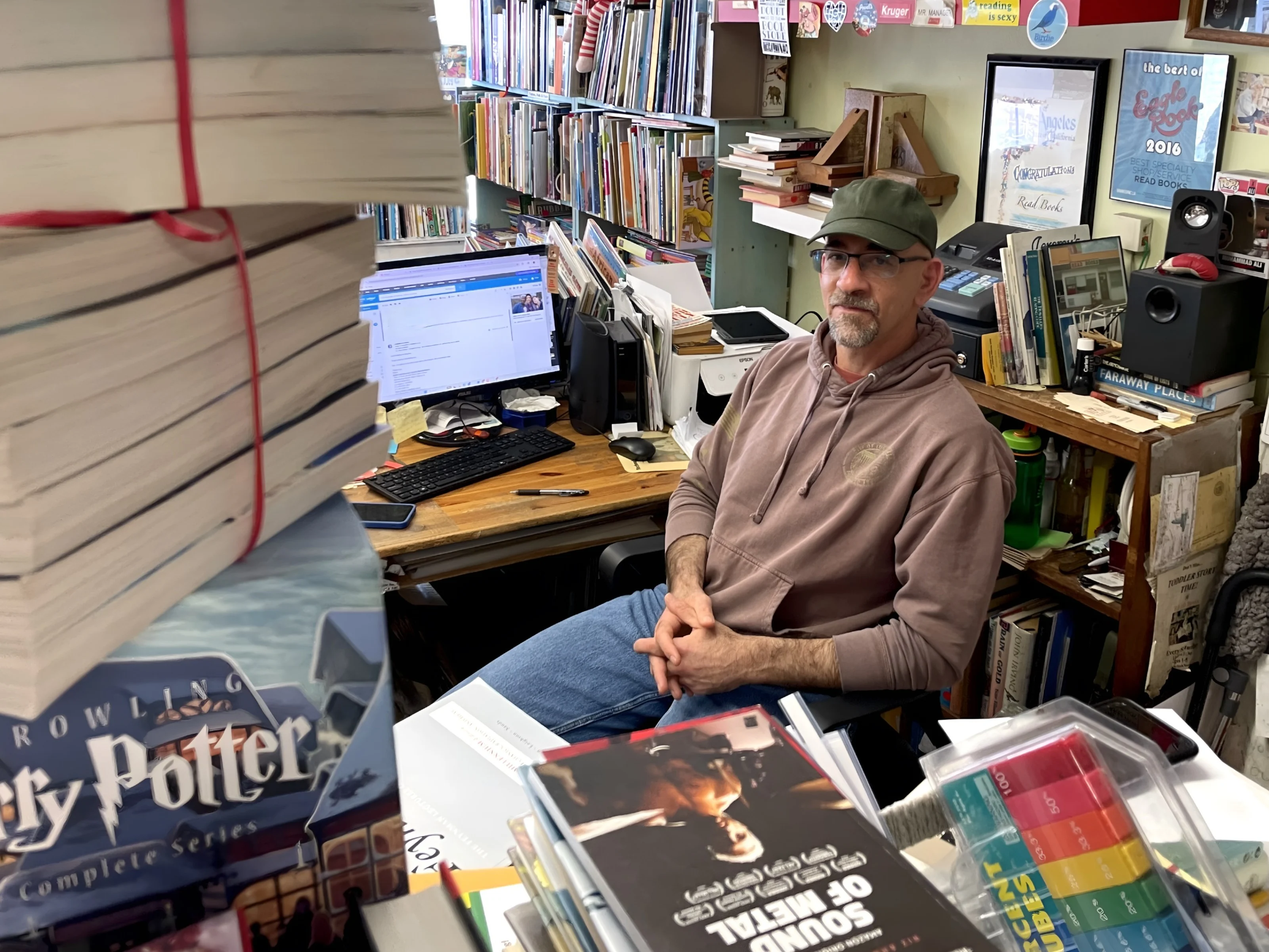 A man sits at a bookstore register surrounded by stacks of books.