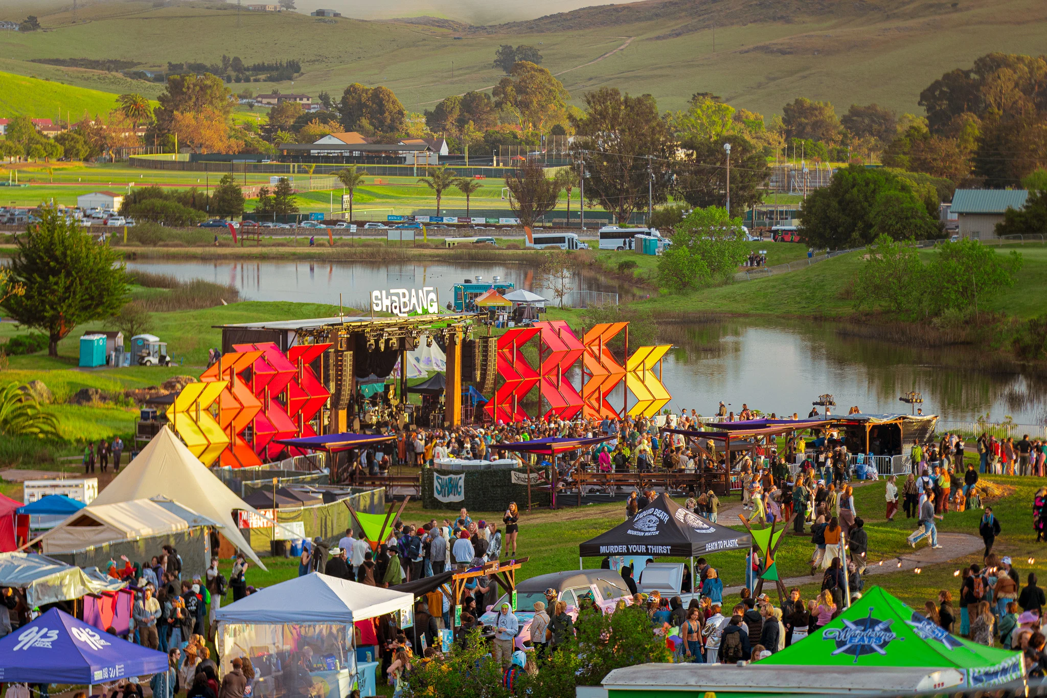 Outdoor music festival with colorful stage decorations beside a lake, surrounded by rolling green hills and crowds of attendees.