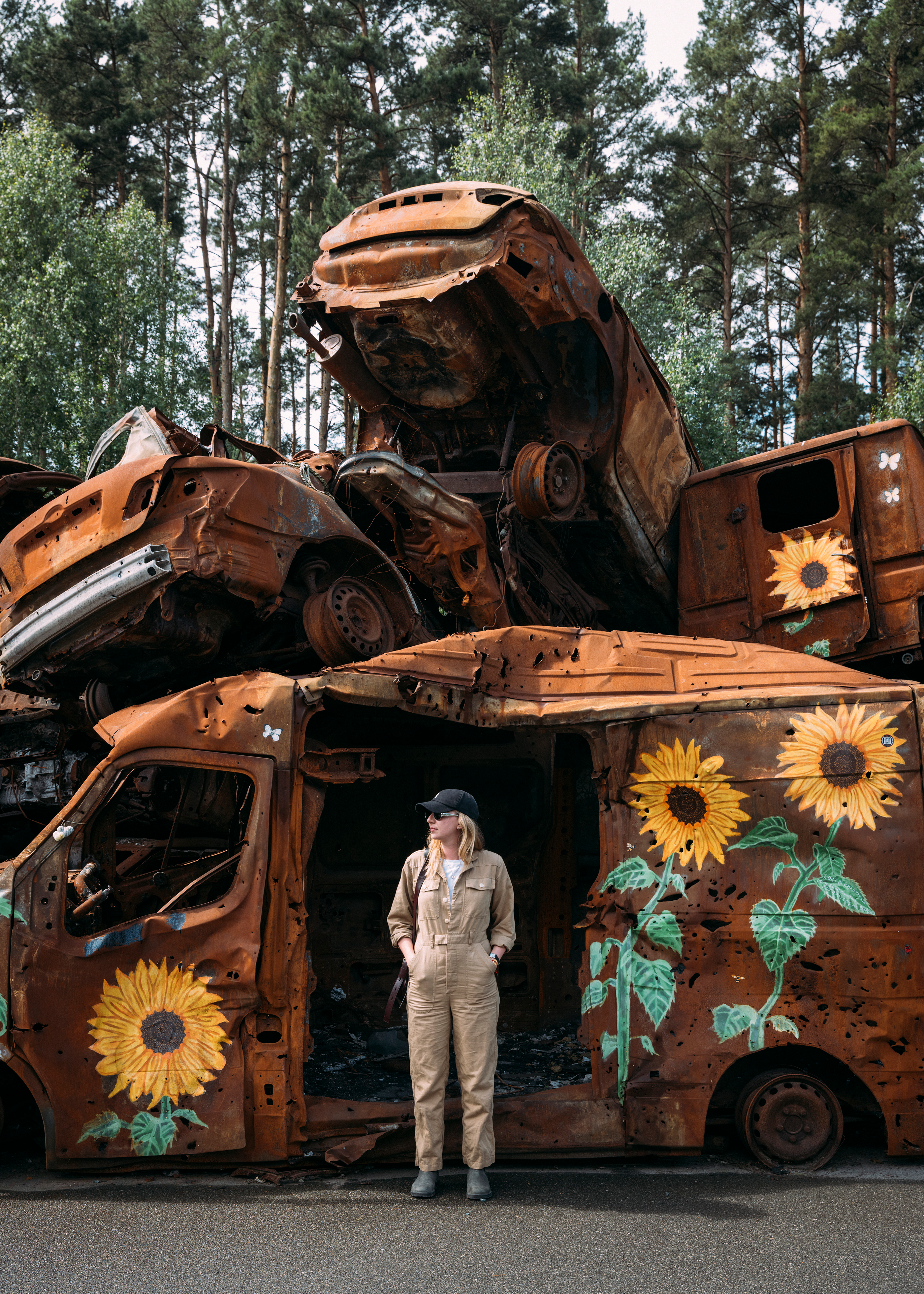 A woman in a tan jumpsuit stands before stacked rusted, war-damaged vehicles painted with sunflowers, set against tall trees.