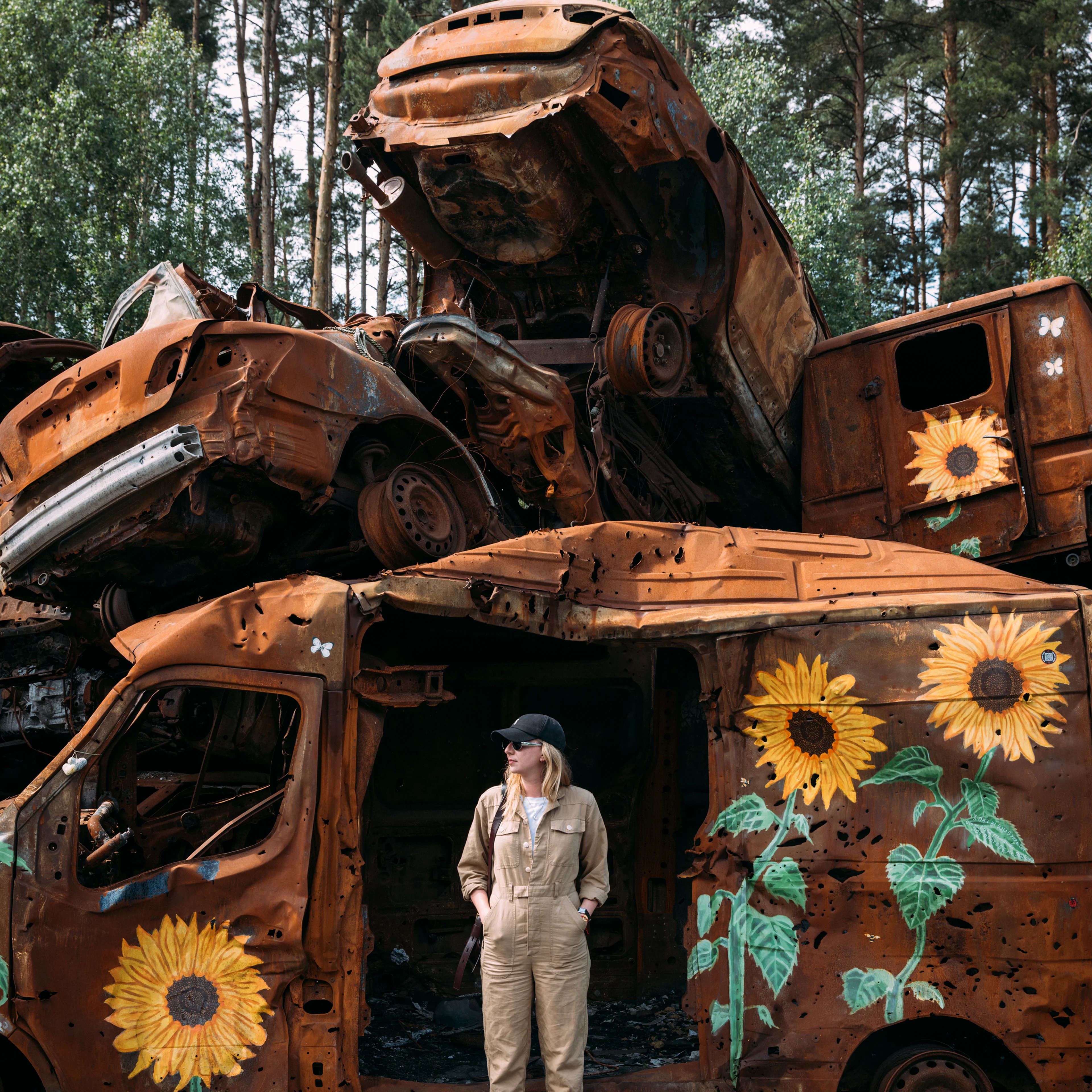 A woman in a tan jumpsuit stands before stacked rusted, war-damaged vehicles painted with sunflowers, set against tall trees.