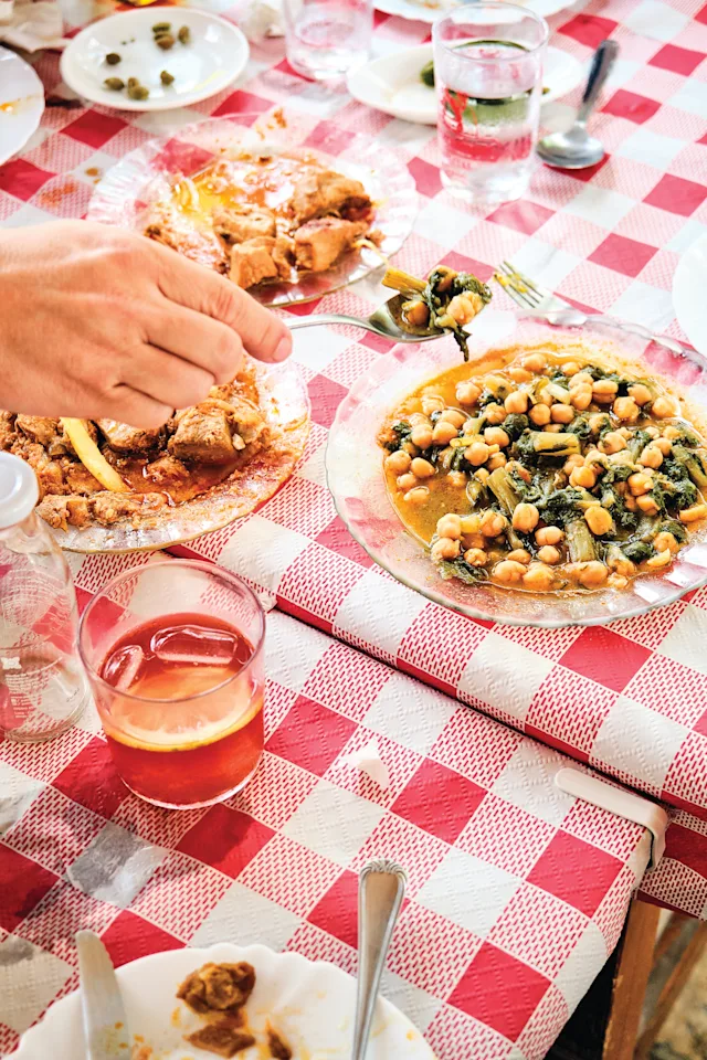 A hand reaches for a plate of meat on a red-checked tablecloth, beside a dish of chickpeas and greens, with drinks nearby.