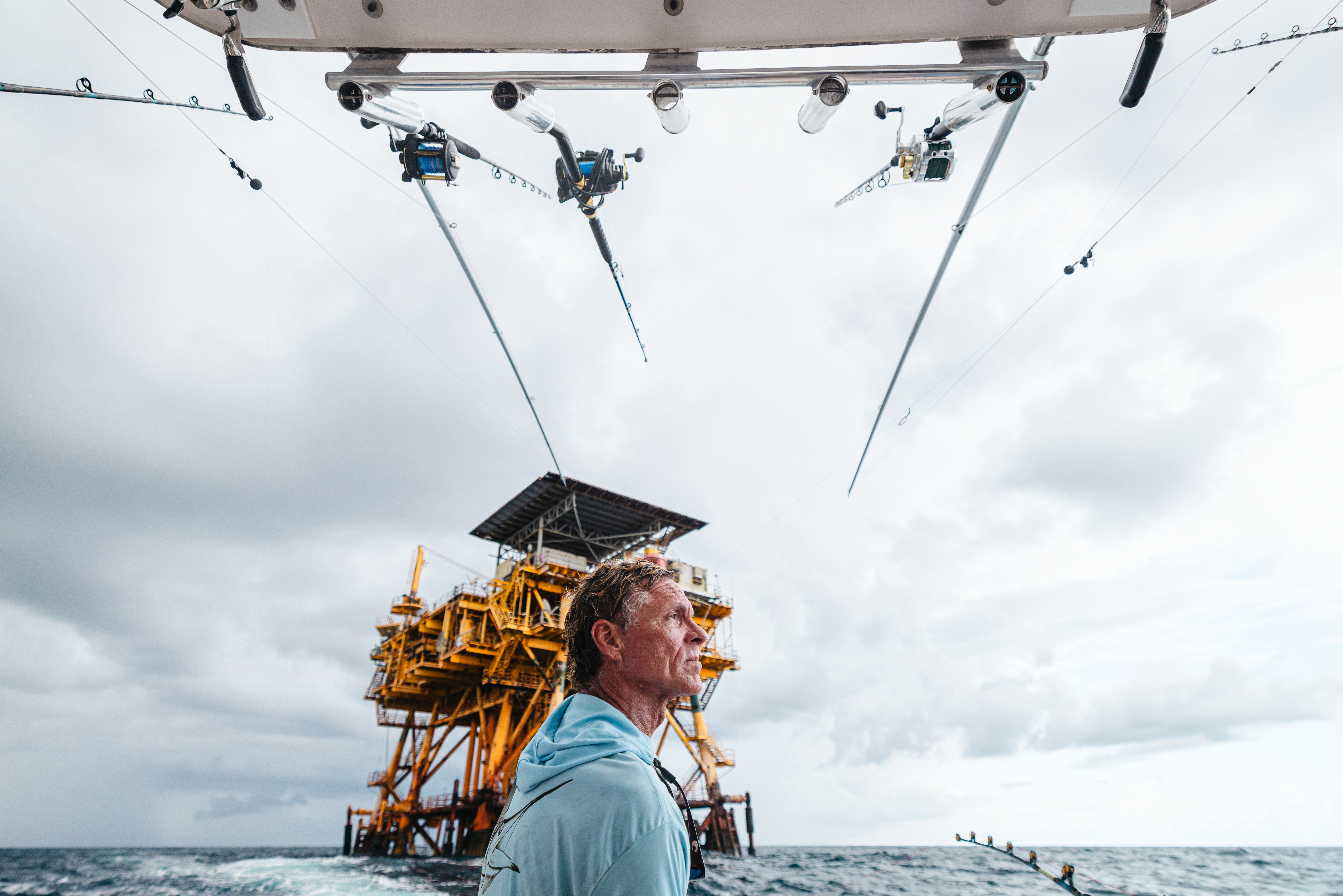 A man in a light blue shirt stands on a fishing boat near an offshore oil platform, with multiple fishing rods overhead.
