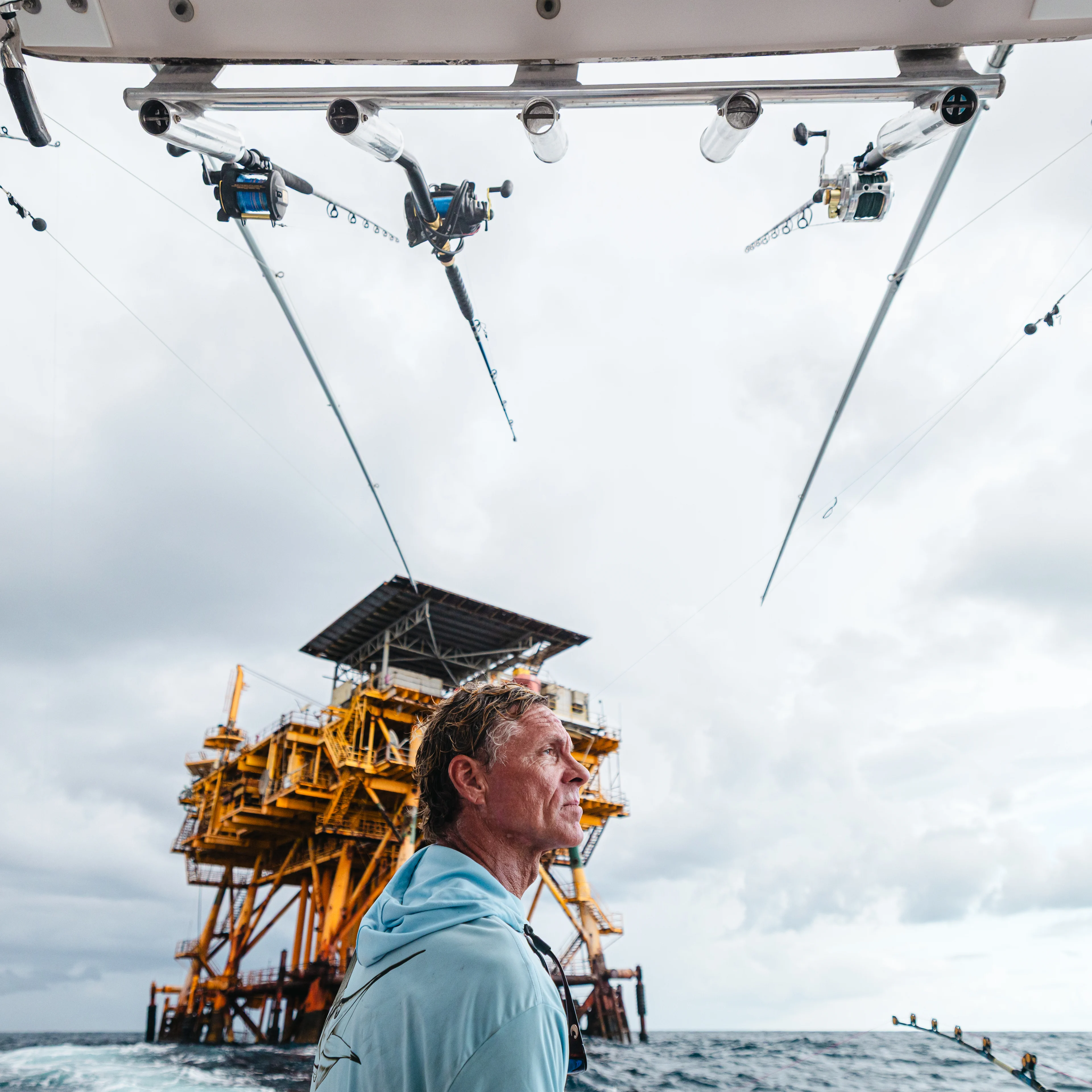 A man in a light blue shirt stands on a fishing boat near an offshore oil platform, with multiple fishing rods overhead.