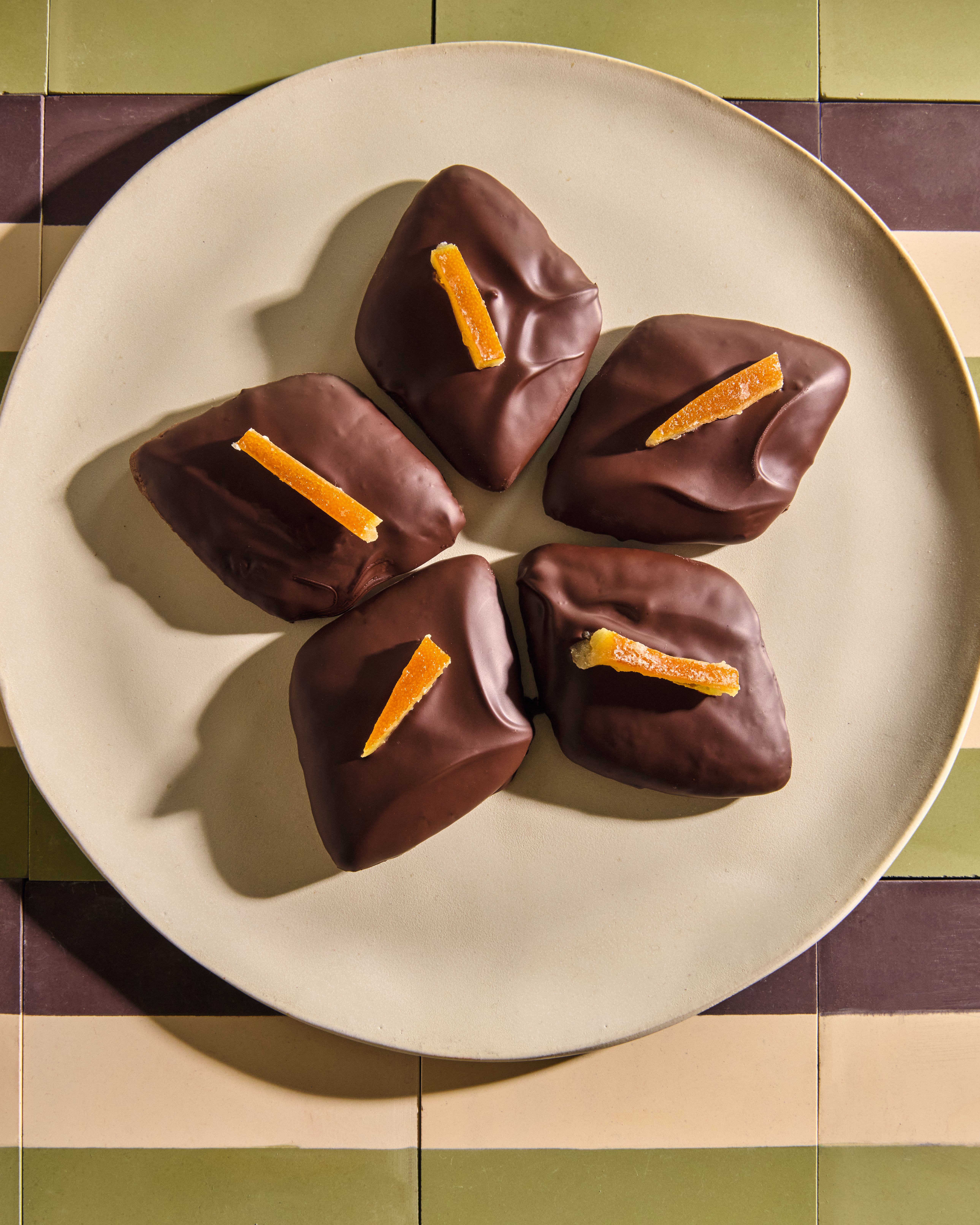 Five dark chocolate-covered treats topped with candied orange peel strips, arranged on a round beige plate.