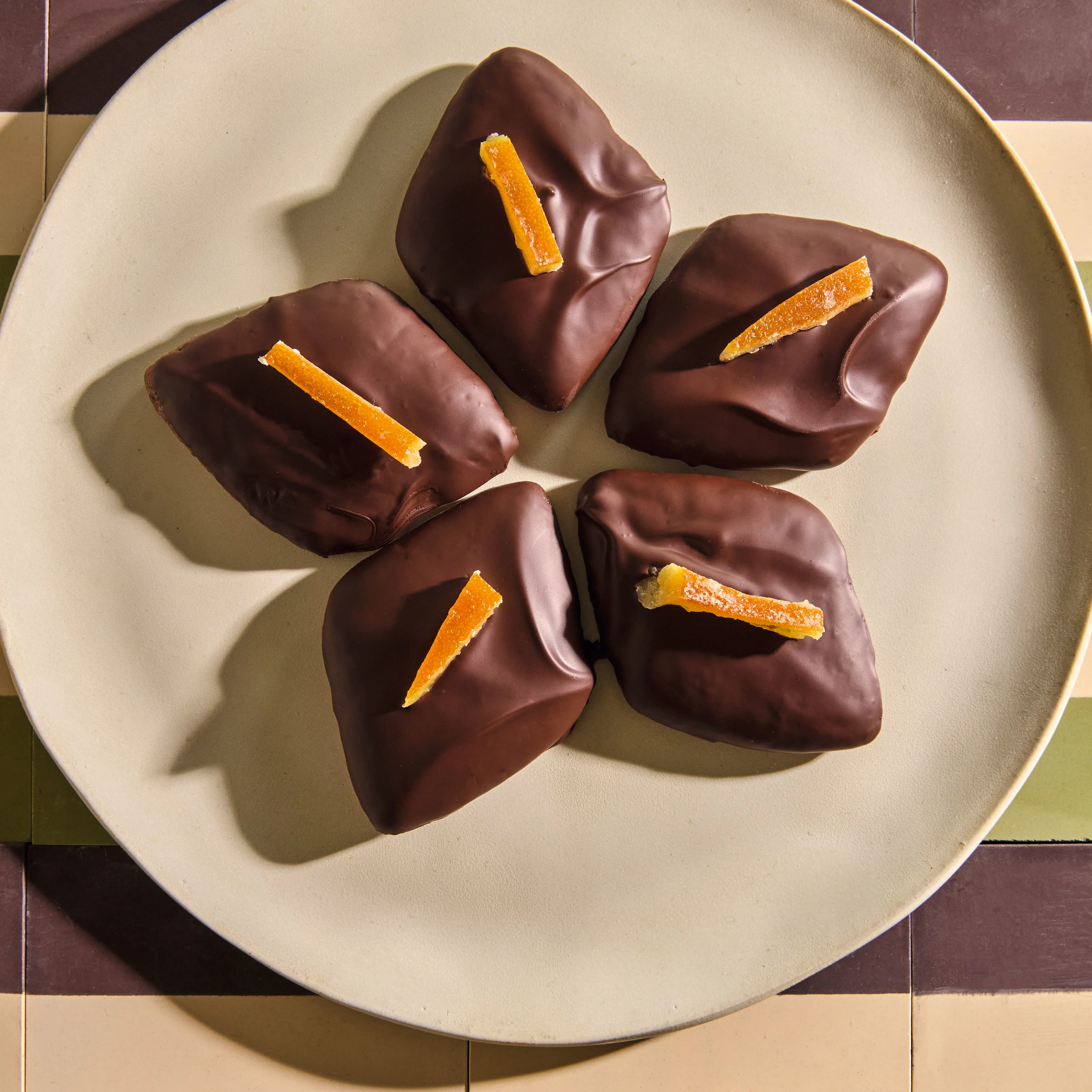 Five dark chocolate-covered treats topped with candied orange peel strips, arranged on a round beige plate.