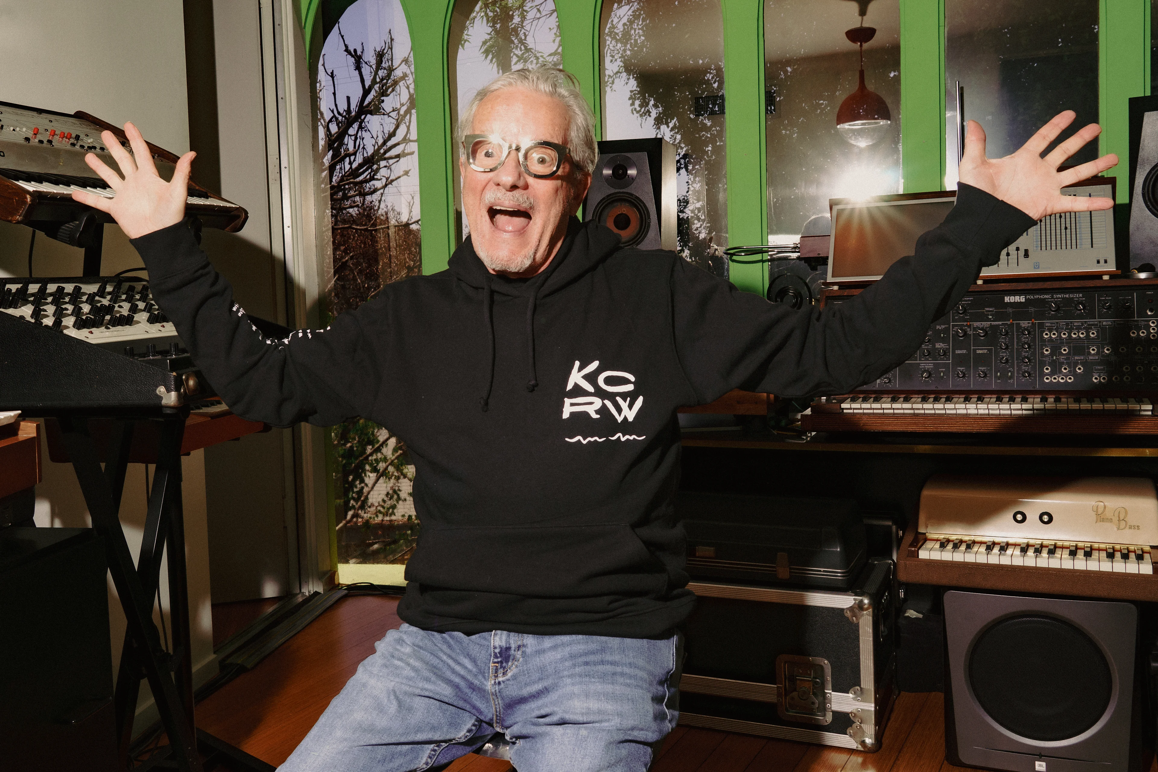 An older man in a black KCRW hoodie raises his hands excitedly in a music studio filled with synthesizers.