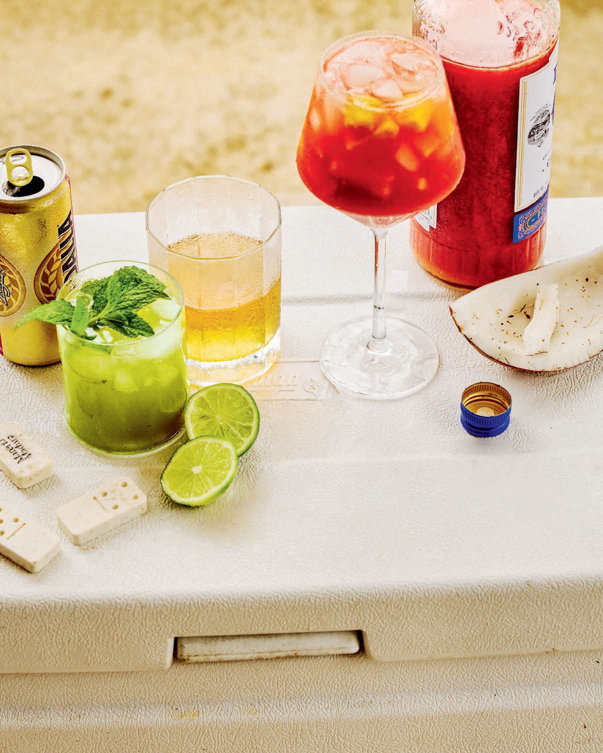 Assorted summer drinks on white cooler including green mojito with lime, beer can, red cocktail, and coconut slice.