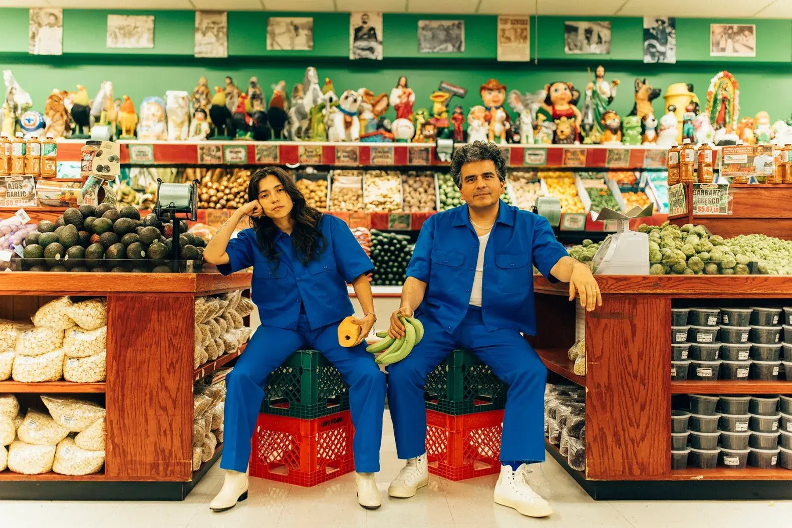 Two people in matching blue jumpsuits sit on crates in a colorful Latin grocery store surrounded by fresh produce and figurines.