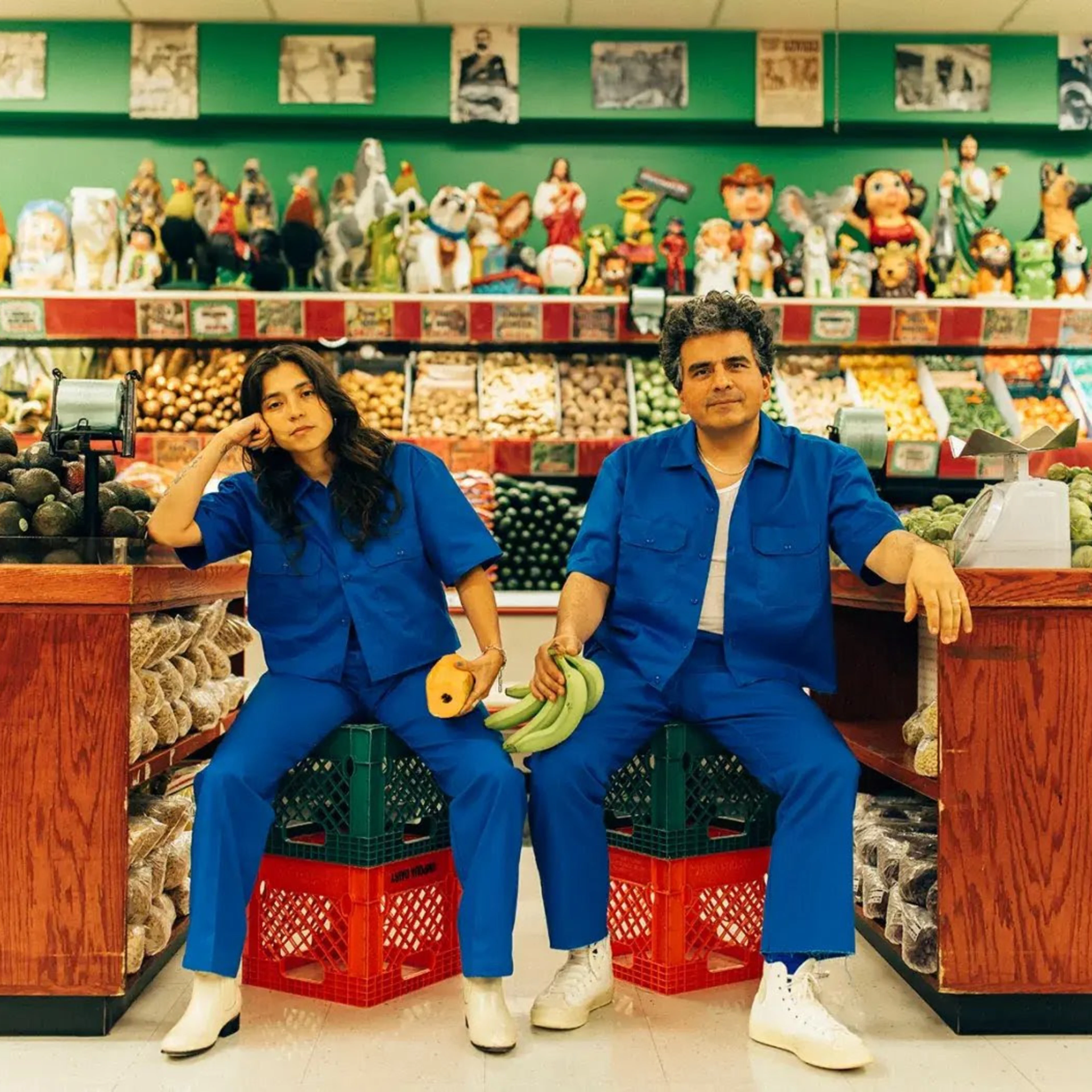 Two people in matching blue jumpsuits sit on crates in a colorful Latin grocery store surrounded by fresh produce and figurines.
