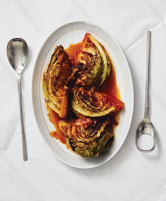 Roasted cabbage wedges in tomato sauce on a white oval platter, flanked by two silver spoons on a white surface.