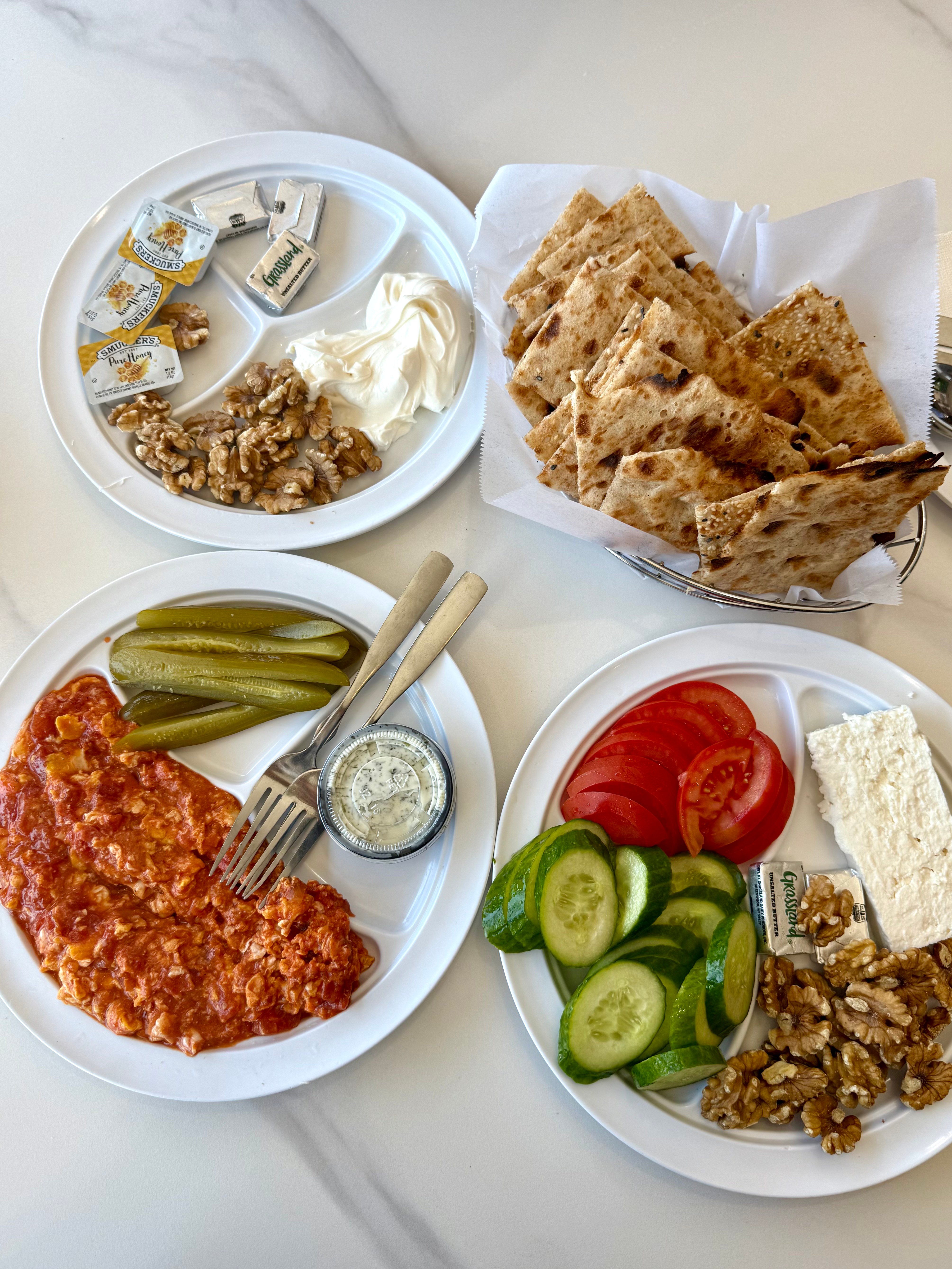 Four white plates with Turkish breakfast items: flatbread, walnuts, cream, tomatoes, cucumbers, feta, pickles, and spiced meat.