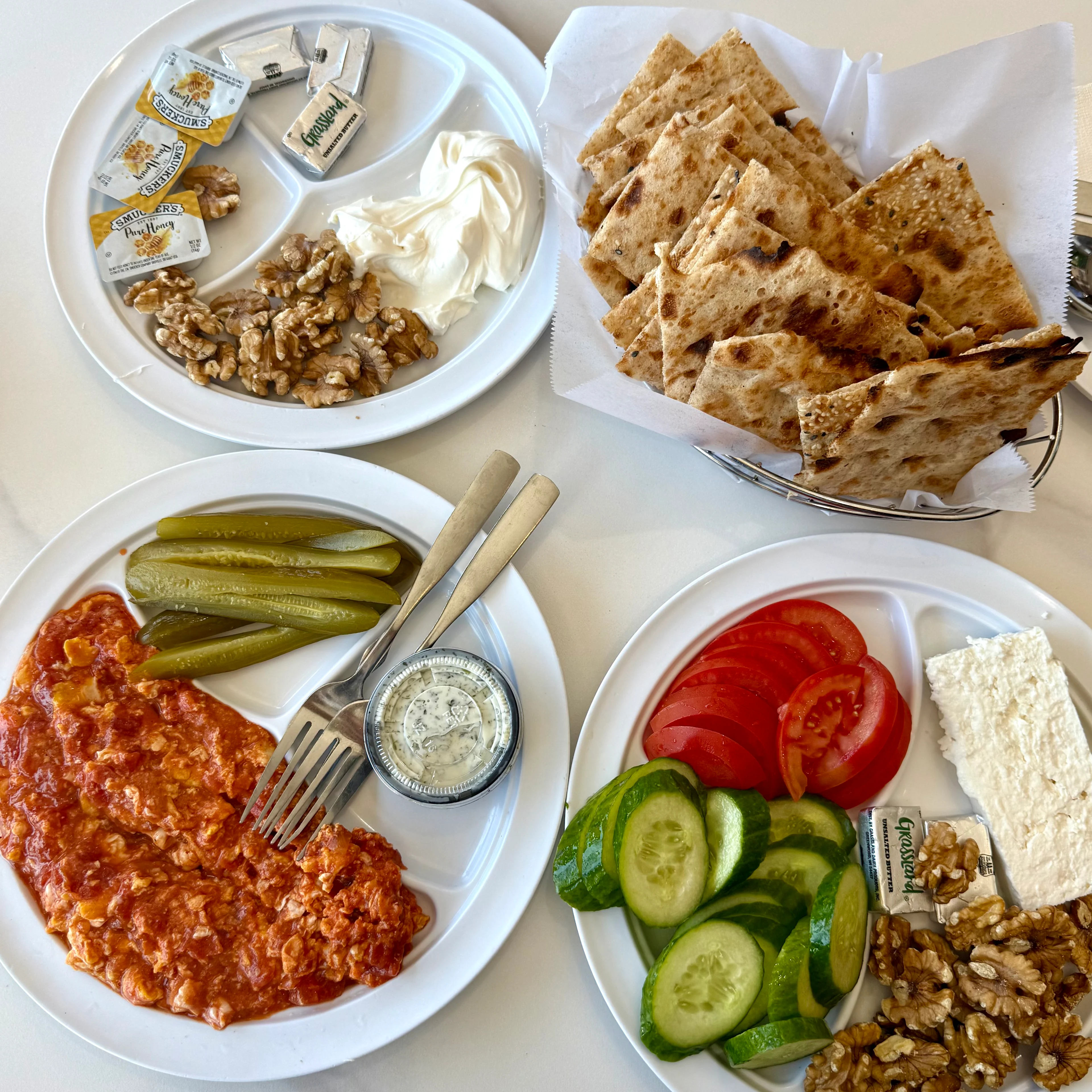Four white plates with Turkish breakfast items: flatbread, walnuts, cream, tomatoes, cucumbers, feta, pickles, and spiced meat.