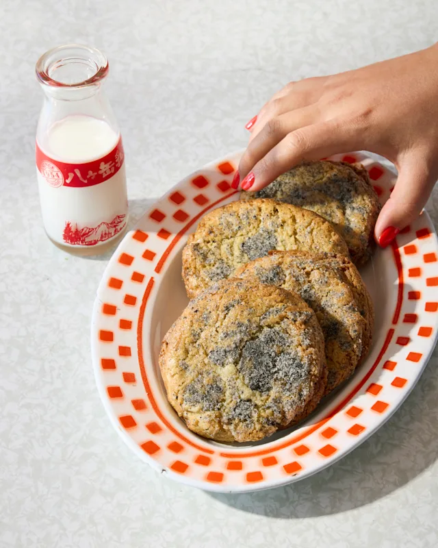 A hand with red nails reaches for cookies on a red-trimmed oval dish, beside a small glass milk bottle.