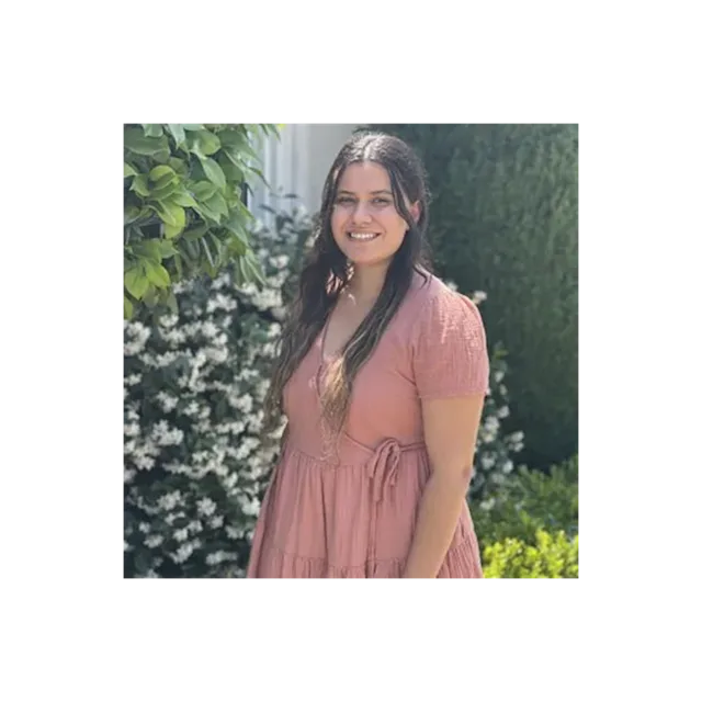 Person in pink dress smiling outdoors with green foliage and white flowering plants in background